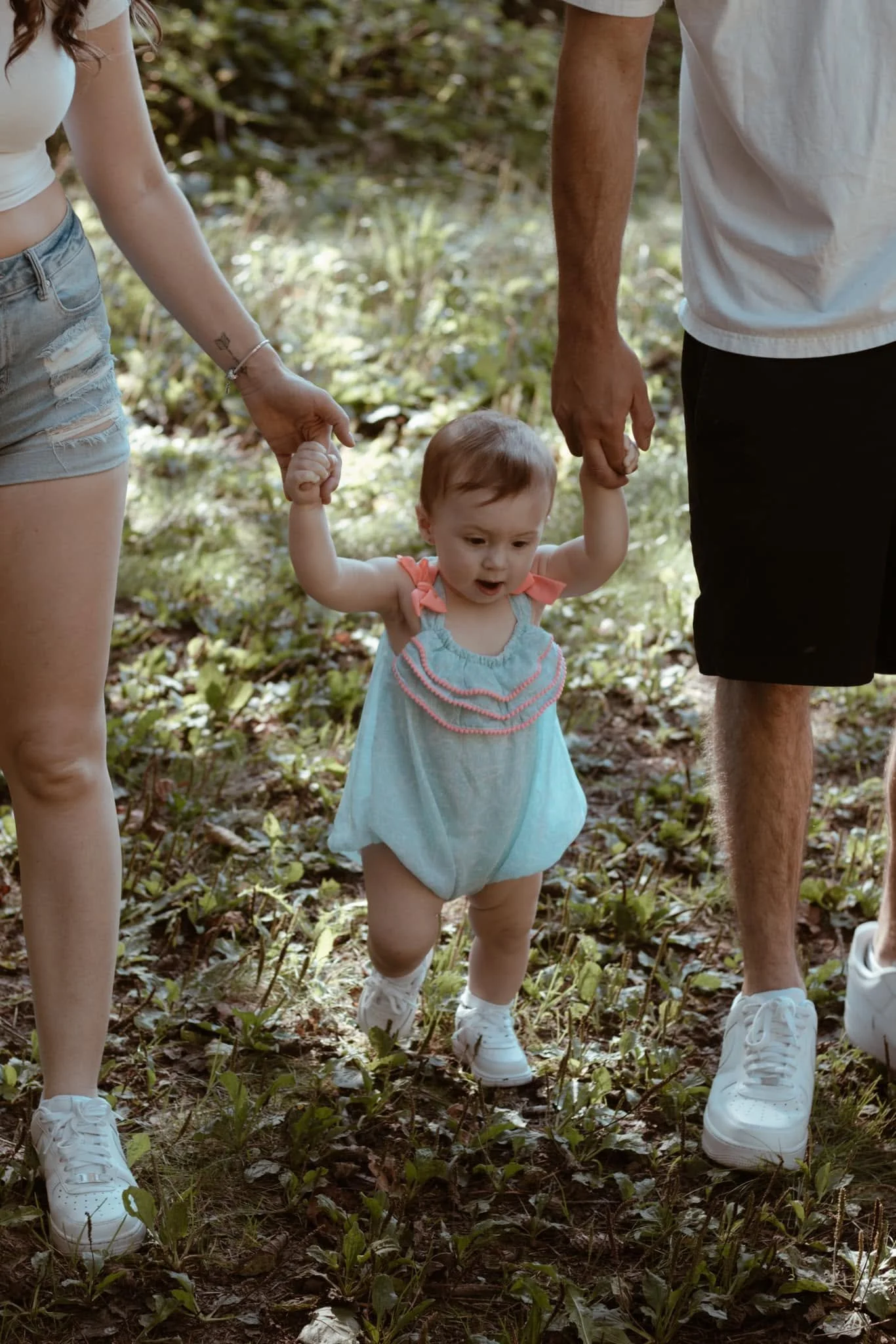 A young girl in a light blue dress walking outdoors between two adults, holding their hands, with a forest floor covered in leaves and patches of grass.