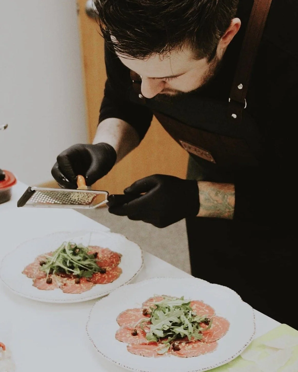 A man wearing black gloves, with dark hair and a tattoo on his arm, is grating cheese over two plates of carpaccio topped with arugula and capers.