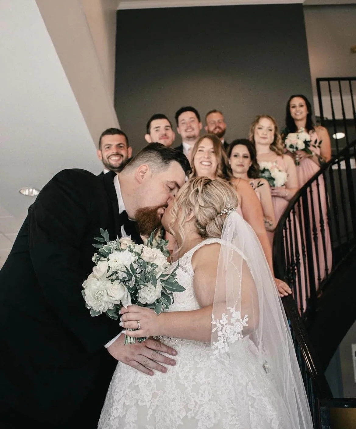 A wedding couple sharing a kiss while holding a bouquet, with friends and family standing on a staircase behind them, smiling.