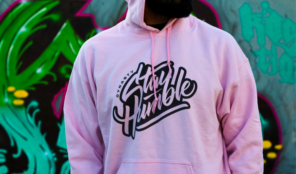 Person wearing a pink hoodie with the words 'Stay Humble' printed on it, standing in front of graffiti art.