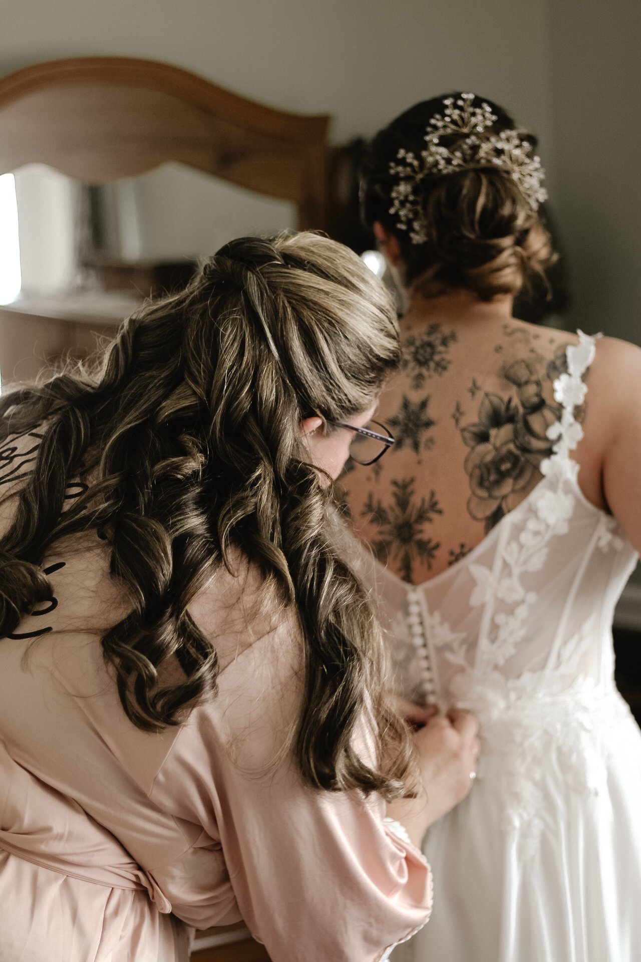 A bride in a lace wedding dress with floral embroidery and a tattoo on her back, standing with her back to the camera. A woman with long, curly hair and glasses, wearing a satin robe, is adjusting or helping the bride.