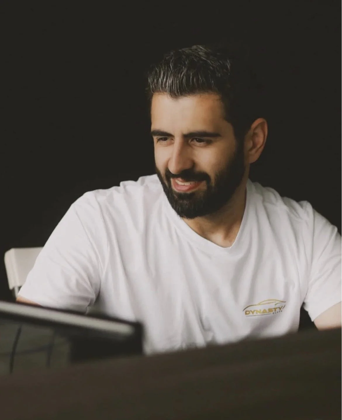 A man with dark hair, beard, and mustache wearing a white shirt looking at a laptop with a smile.