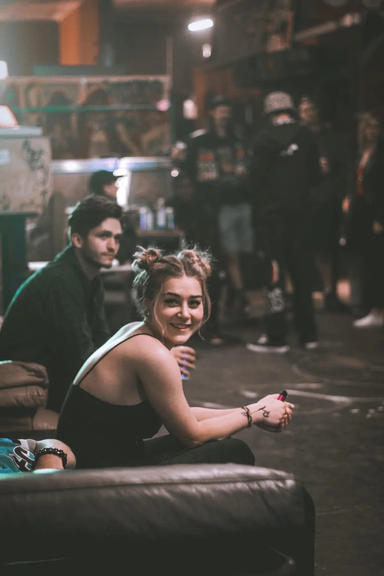 A young woman with red hair styled in buns smiling and sitting on the floor, holding a small object in her hands, in a dimly lit indoor setting with people in the background.