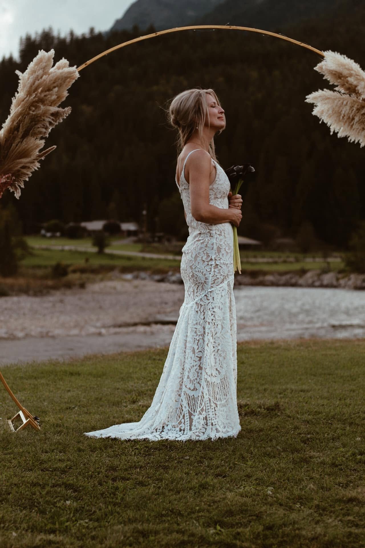 A woman in a lace white dress standing outdoors with eyes closed, holding a dark flower, framed by pampas grass, mountain and trees in the background.