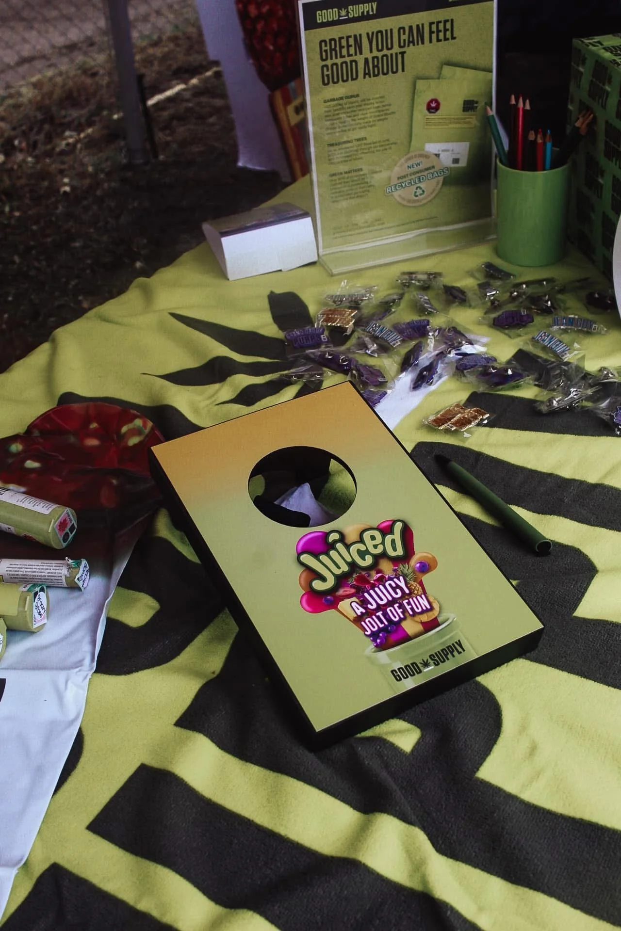 A display table with a yellow and black striped cloth, featuring a Juiced energy drink box, small candy bags, pens, and promotional posters about green cannabis use and recycled bags, at an outdoor event.