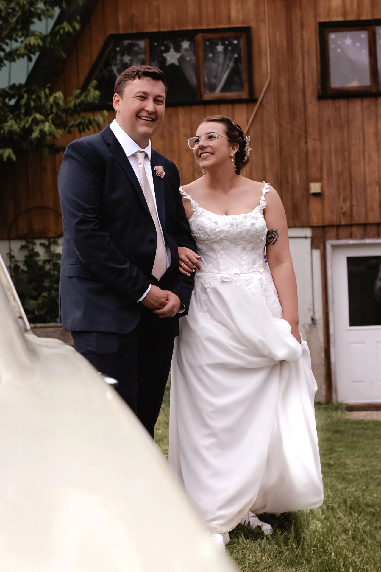 A smiling man in a dark suit and a smiling woman in a white wedding dress standing outdoors in front of a wooden building with star decorations in the windows.