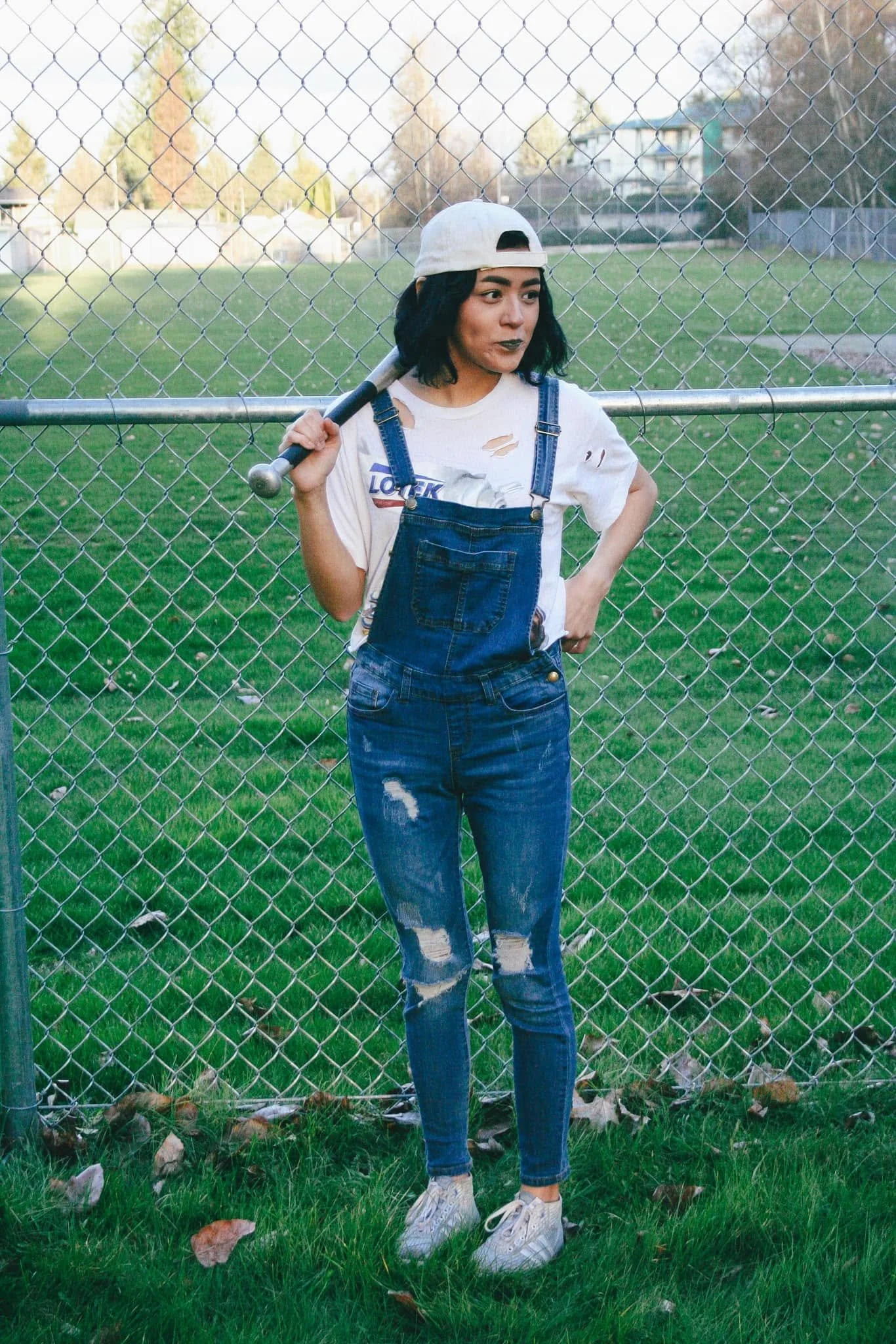 Young woman with black hair wearing a white baseball cap, ripped white T-shirt, denim overalls, and white sneakers, standing in front of a chain-link fence on a grassy field, holding a baseball bat on her shoulder, with a suburban neighborhood in the