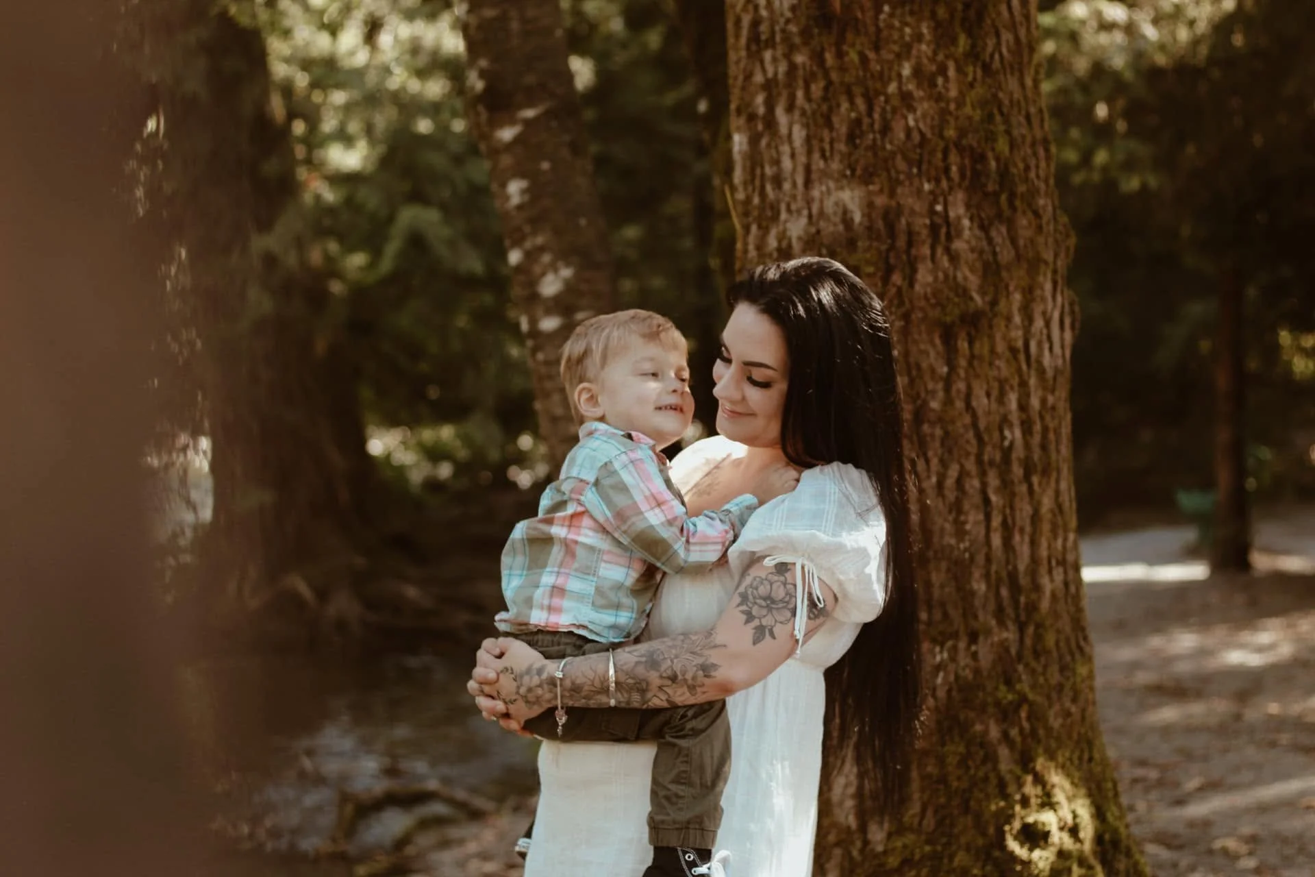 A woman with black hair and tattoos holds a young boy with red hair in a forest with tall trees, smiling at each other.