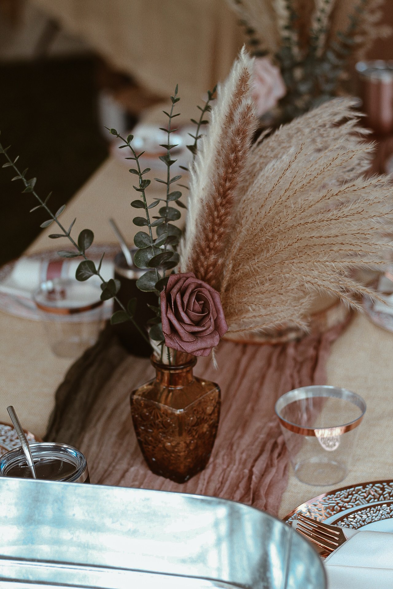 A floral arrangement in a small amber glass vase, featuring a single pink rose, eucalyptus leaves, and pampas grass, placed on a wooden decorative piece on a table.