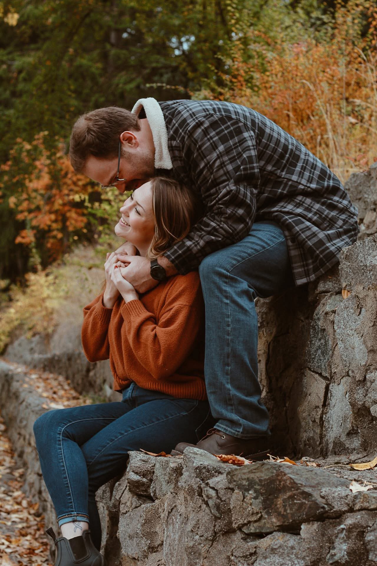 A man and woman sharing a joyful moment outdoors during fall, with the man bending over to kiss the woman on the head while she smiles happily, sitting on a stone wall surrounded by autumn trees.