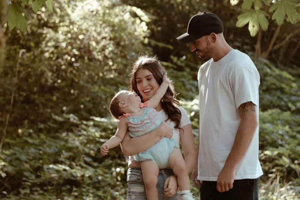 A family of three outdoors, a woman holding a young girl and a man standing nearby, all smiling and enjoying time together in a wooded area.