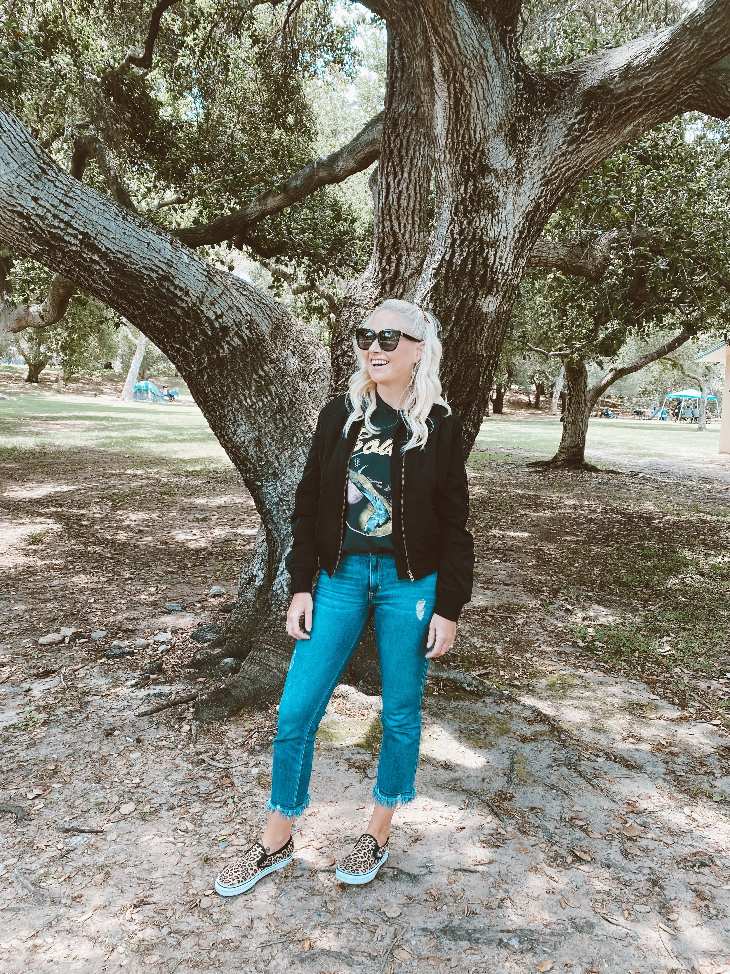 A woman standing outdoors next to a large tree, smiling and wearing sunglasses, black jacket, graphic t-shirt, ripped jeans, and leopard print slip-on shoes.