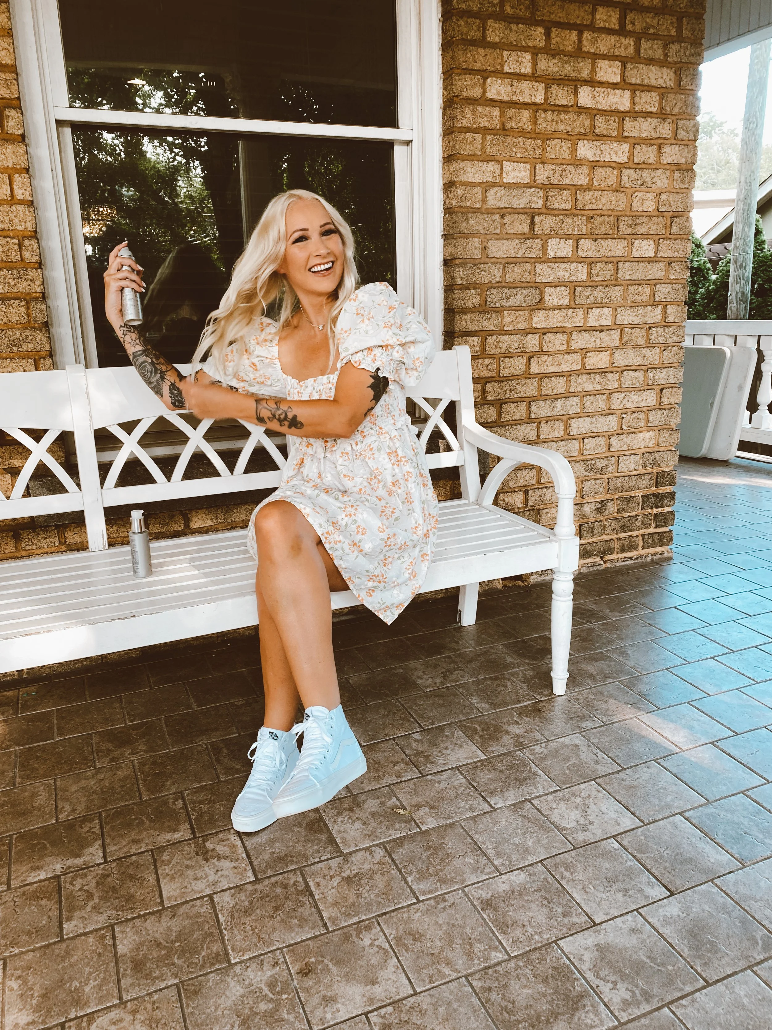 A woman with long blonde hair, wearing a floral dress and white sneakers, sits on a white garden bench outside a brick house. She is smiling widely and spraying perfume from a can. There are two small bottles on the bench.