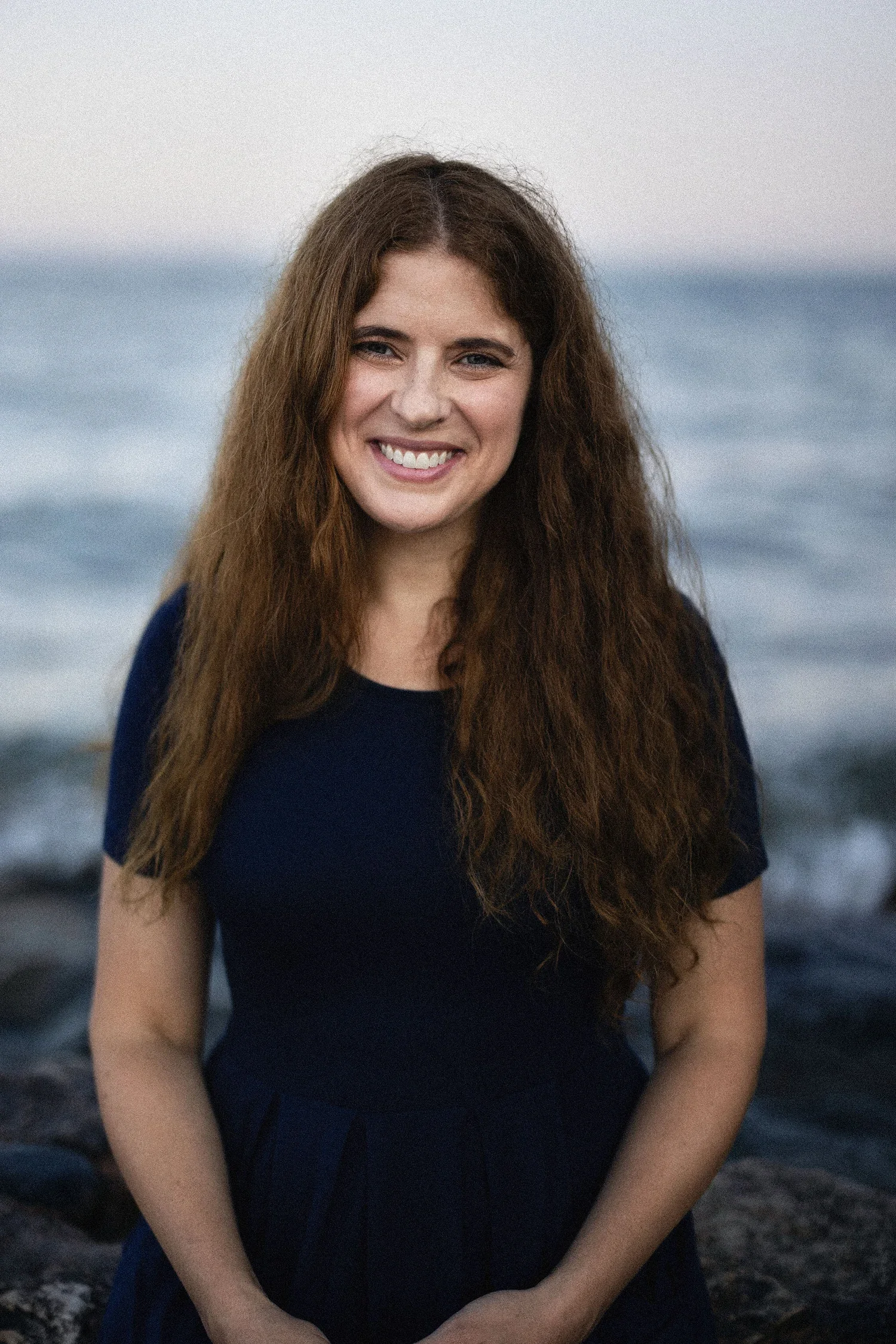 A young woman with long, curly red hair smiling at the camera while standing outdoors near the ocean, wearing a dark blue top.