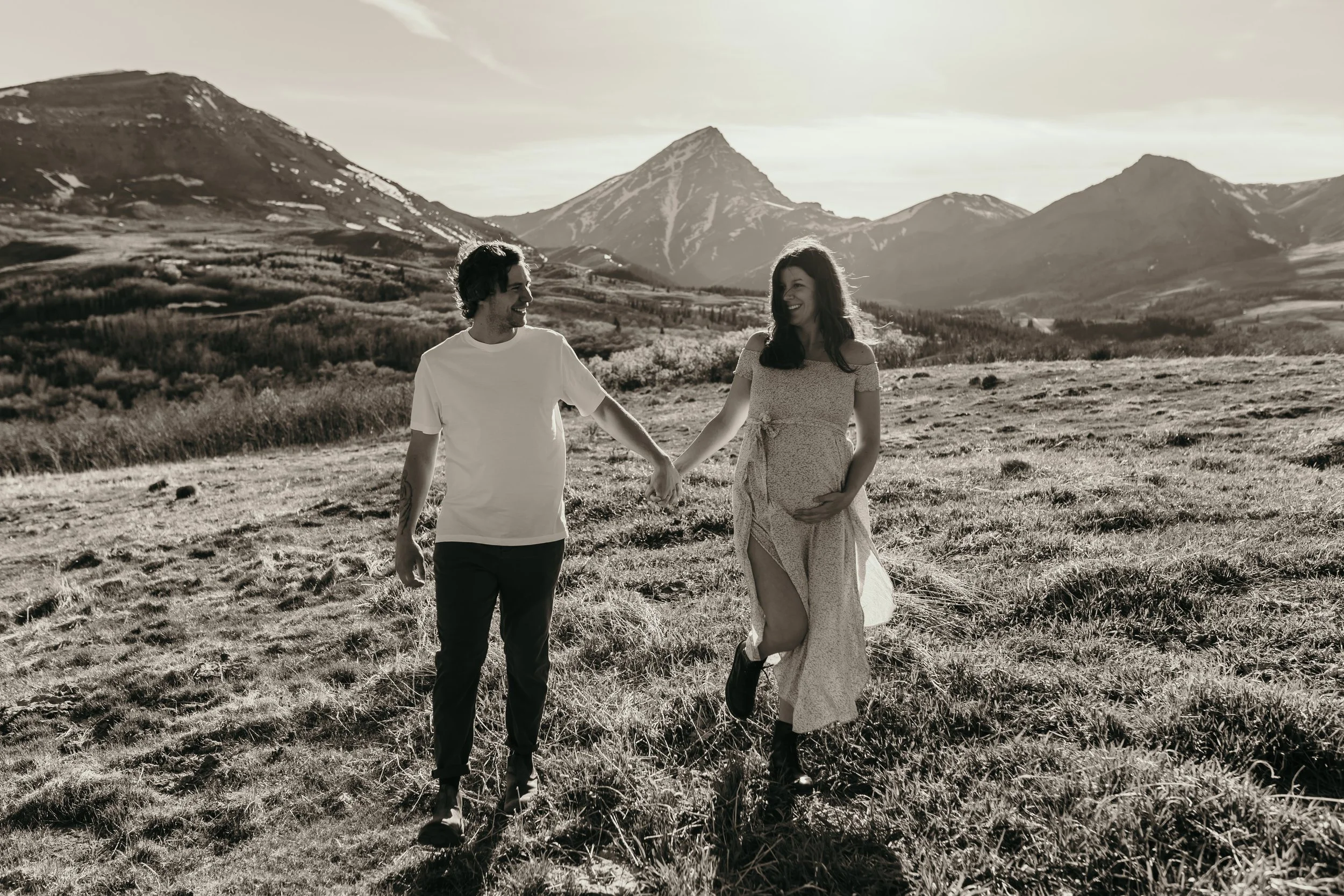 A smiling couple holding hands while walking in a grassy field with mountains in the background, in black and white.