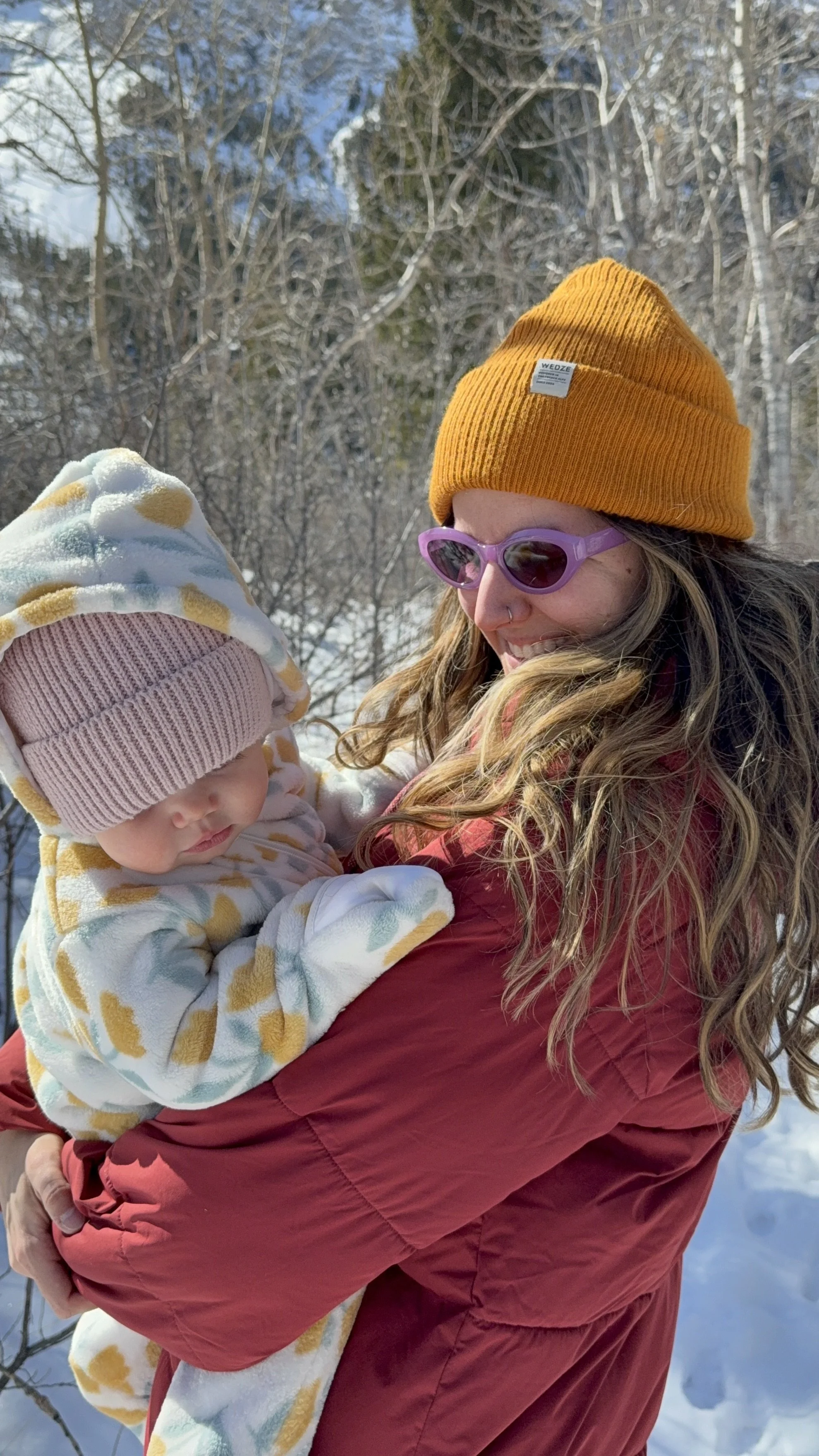 A woman wearing an orange beanie and purple sunglasses is holding a baby dressed in a patterned jacket and pink hat outdoors in a snowy landscape.