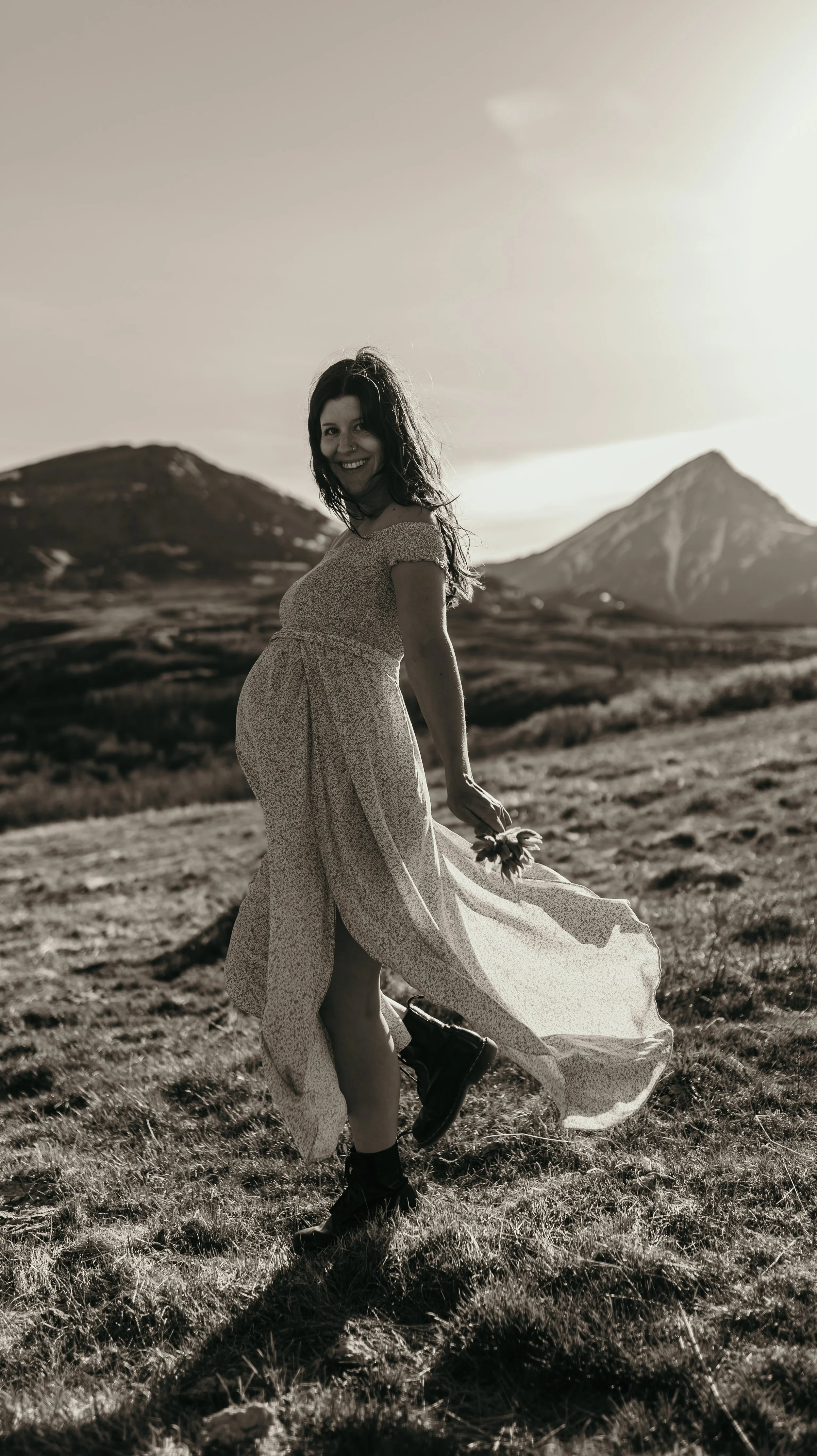 A healthy pregnant woman in a flowy dress holding a flower, smiling, walking in an open field at sunset with mountains in the background.