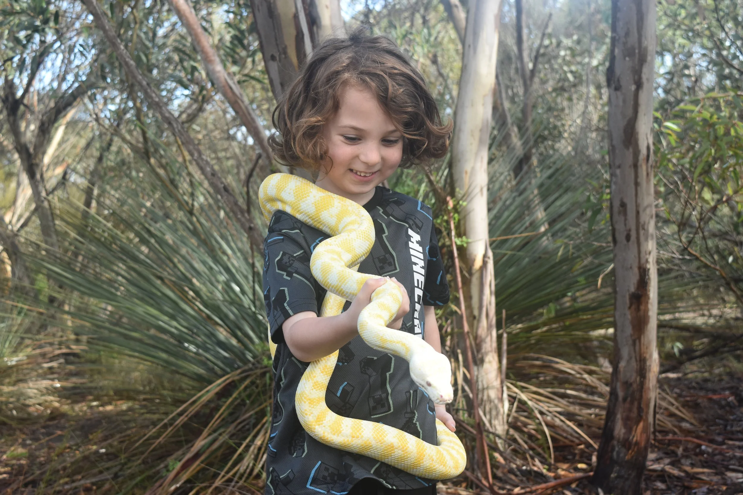 A young boy with curly hair smiling in a wooded area, holding a yellow and white snake.