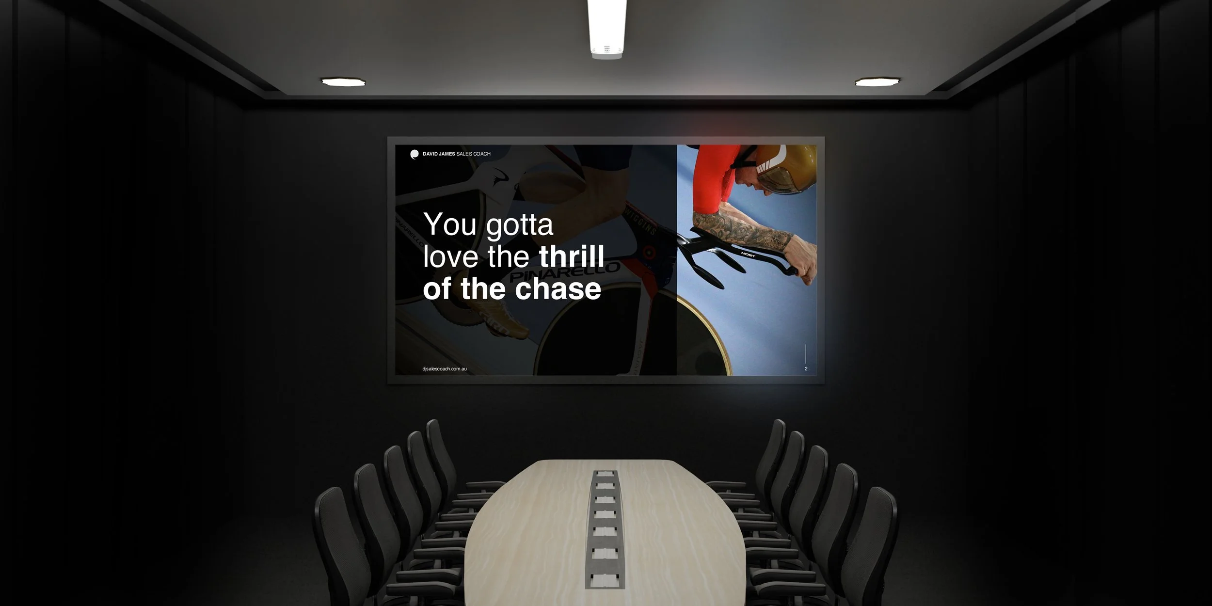 A conference room with a long table and nine black chairs, black walls, ceiling lights, and a large poster on the front wall showing a cyclist in red and black gear riding a bike with the text 'You gotta love the thrill of the chase'.