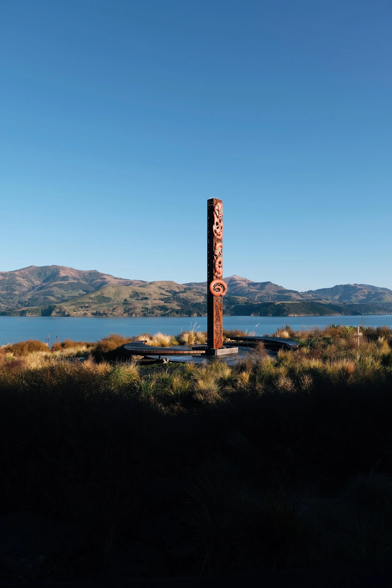 Takapuneke, Akaroa. A tall, carved totem pole stands in a field of grass near a body of water, with mountains in the background and a clear blue sky above.Photography by Julia Atkinson-Dunn