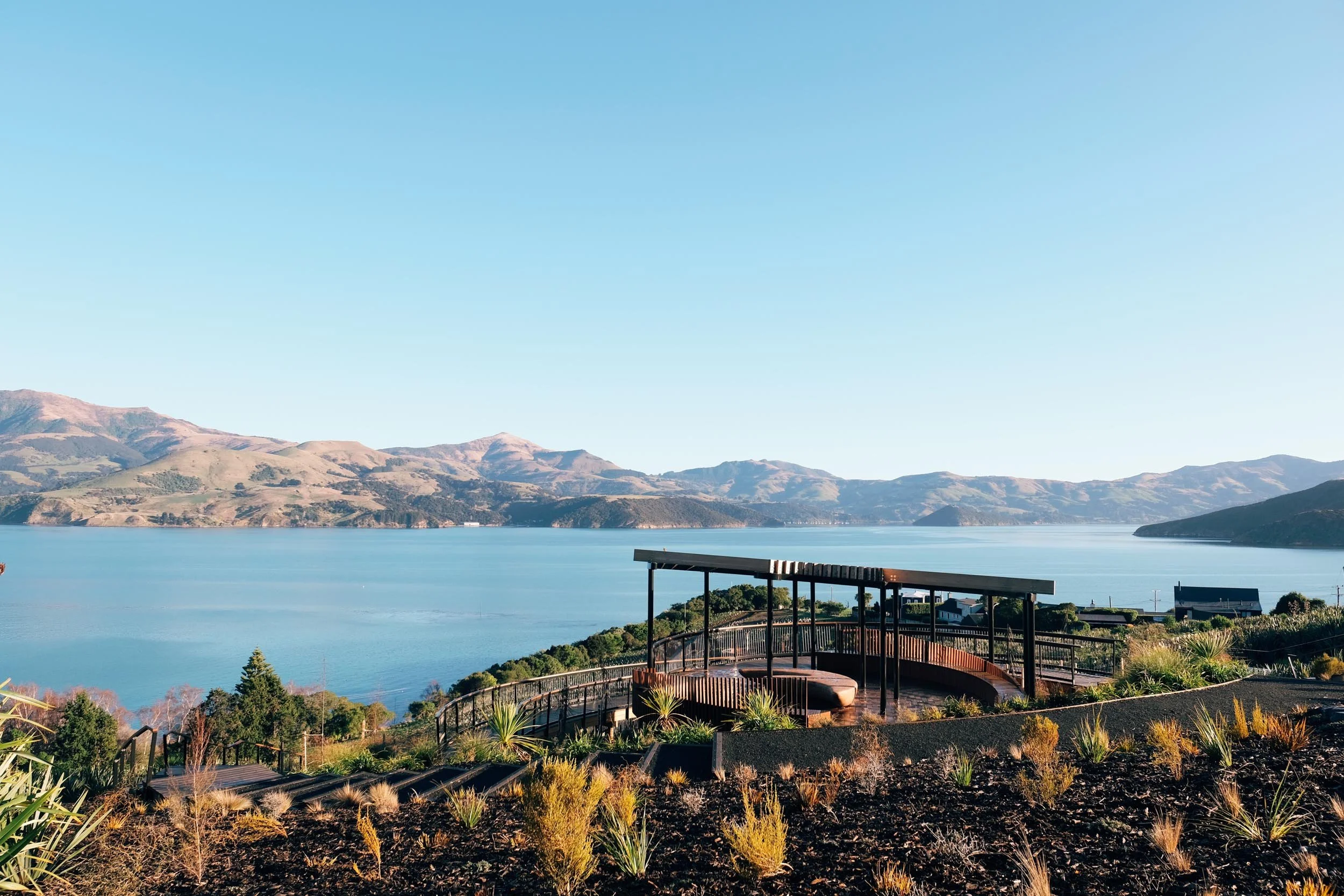 Takapuneke, Akaroa. Scenic view of a lake with mountains in the background and a wooden observation deck in the foreground on a clear, sunny day. Photography by Julia Atkinson-Dunn