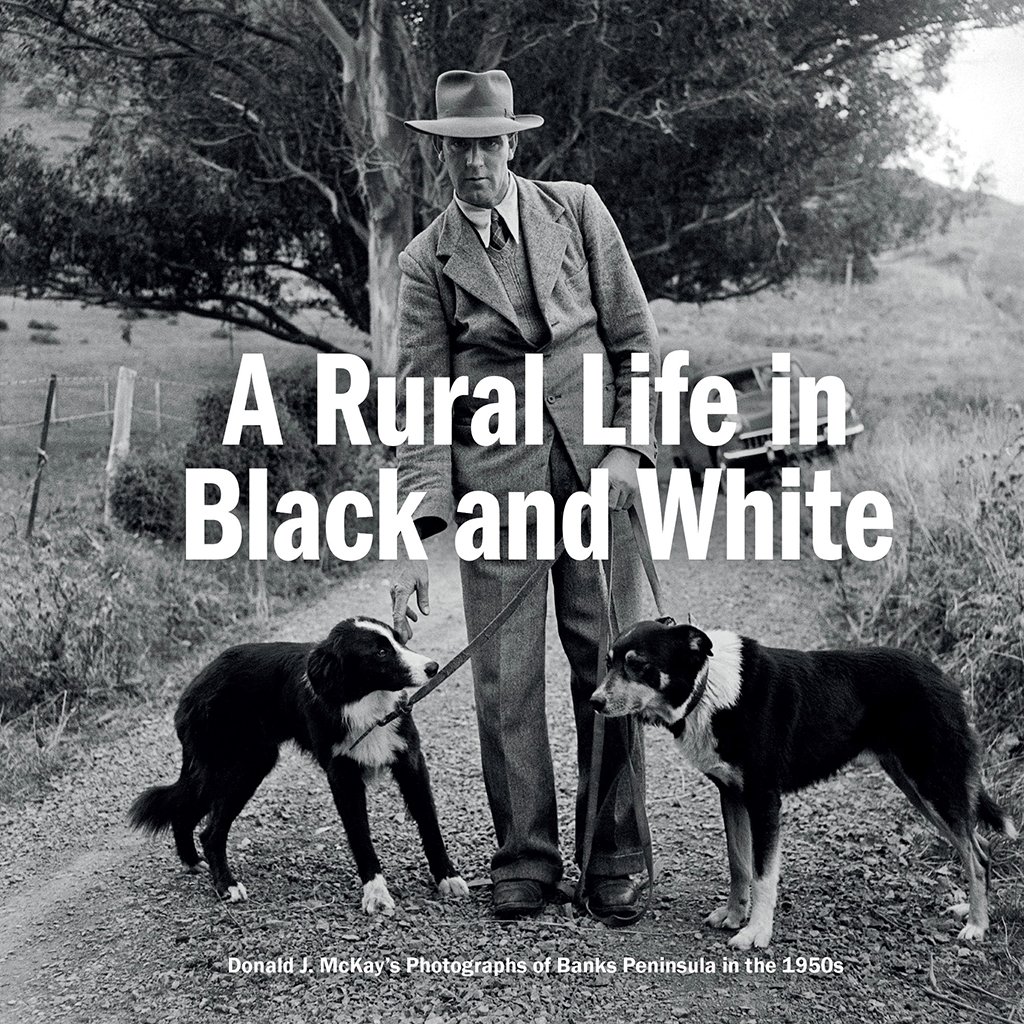 A black and white photo of a man in vintage attire from the 1950s standing on a dirt path with two dogs, in a rural landscape with trees and open fields, with the text "A Rural Life in Black and White" overlaid. Produced by Akaroa Museum