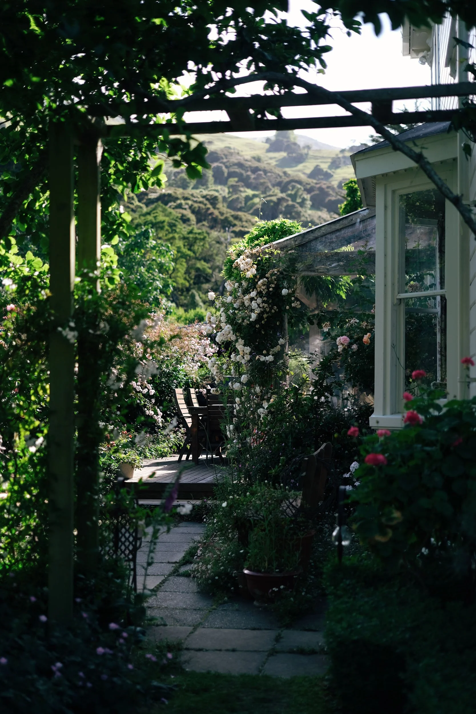 A lush garden with a pathway leading to an outdoor sitting area, surrounded by blooming flowers and greenery, with a house on the right and hills in the background.