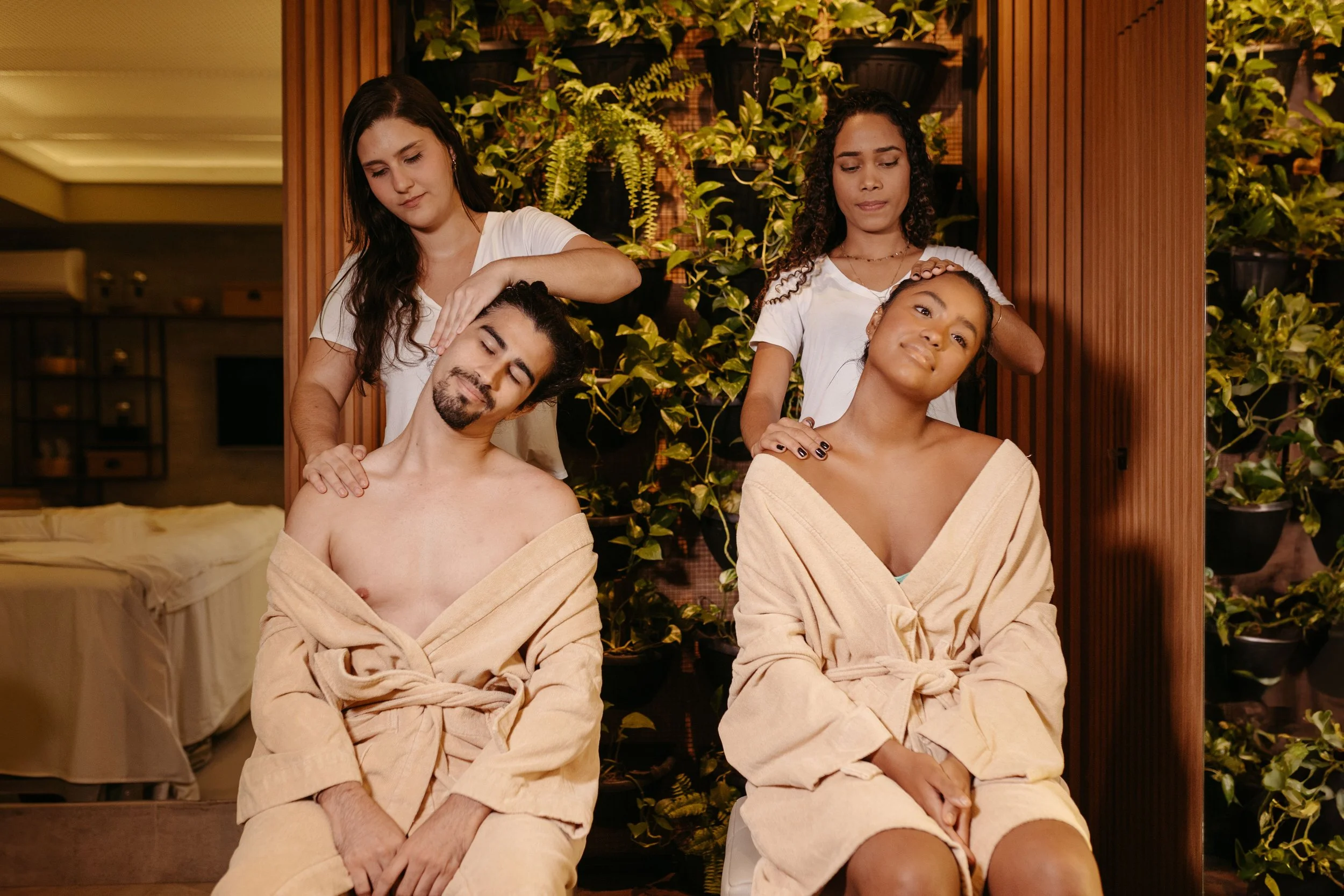 Two women giving massages to a man and a woman who are seated with eyes closed, wearing towels, in a spa or wellness center with green plants in the background.