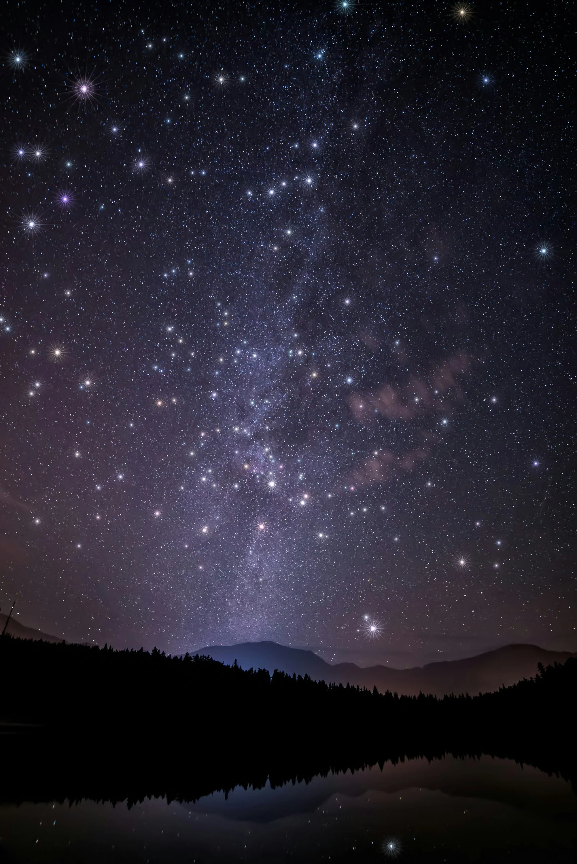 Night sky filled with stars, including the bright constellation of the Pleiades star cluster, over a silhouette of mountains and a calm lake reflecting the starry sky.