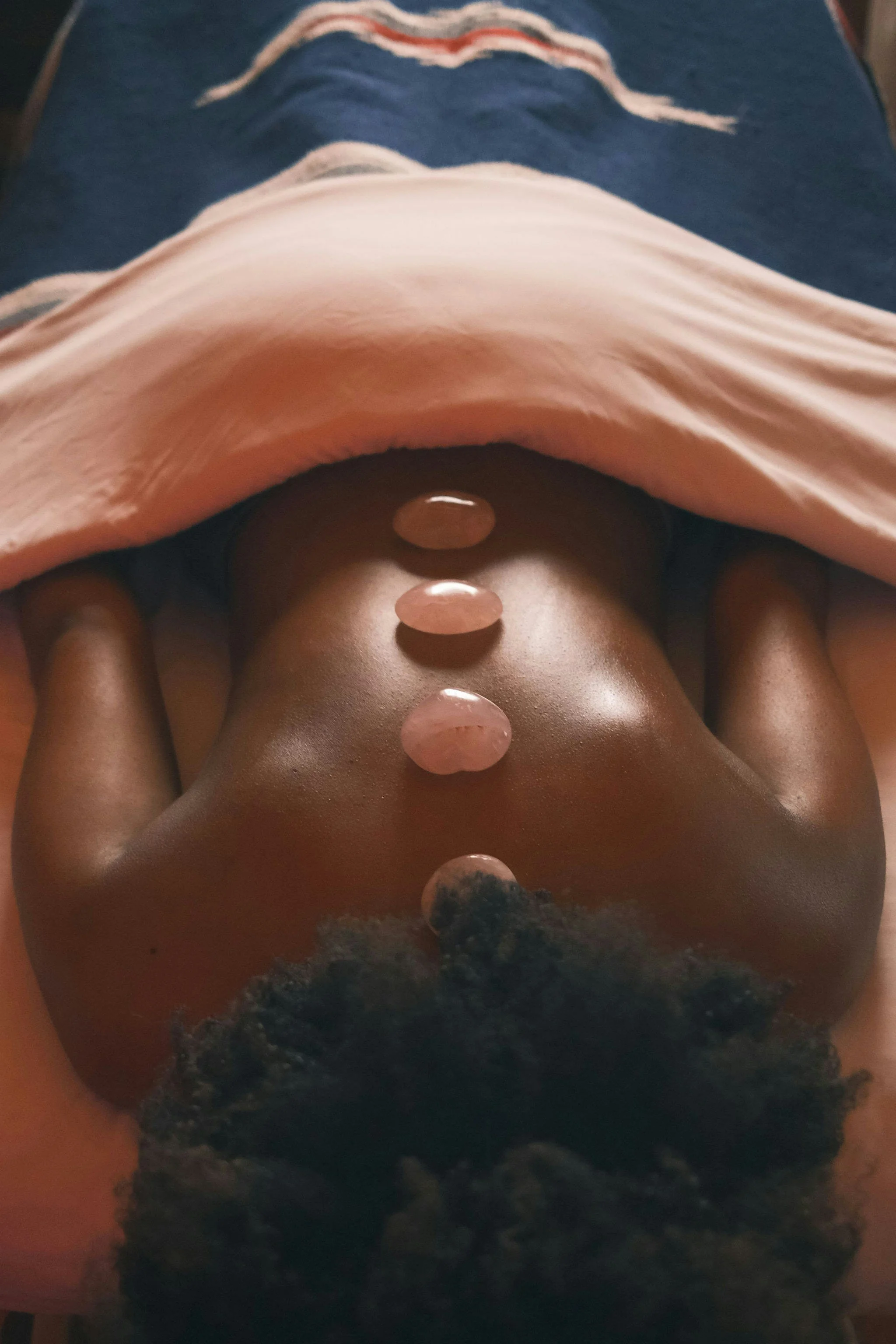 A person lying on their back with six rose quartz stones placed along their body from the chest to the lower abdomen. The person is wearing a peach-colored top and dark shorts, with a dark curly hairstyle. The background features a blue blanket with a red, white, and blue design.