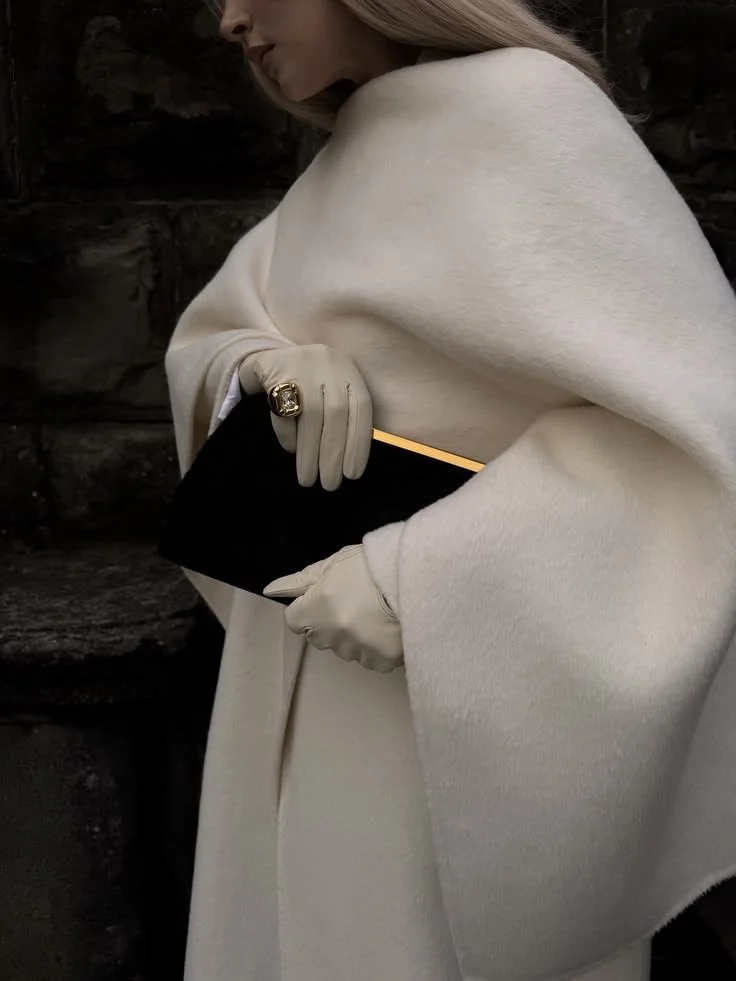 A woman dressed in a cream-colored coat and gloves holding a black clutch purse with a gold-embellished pen.