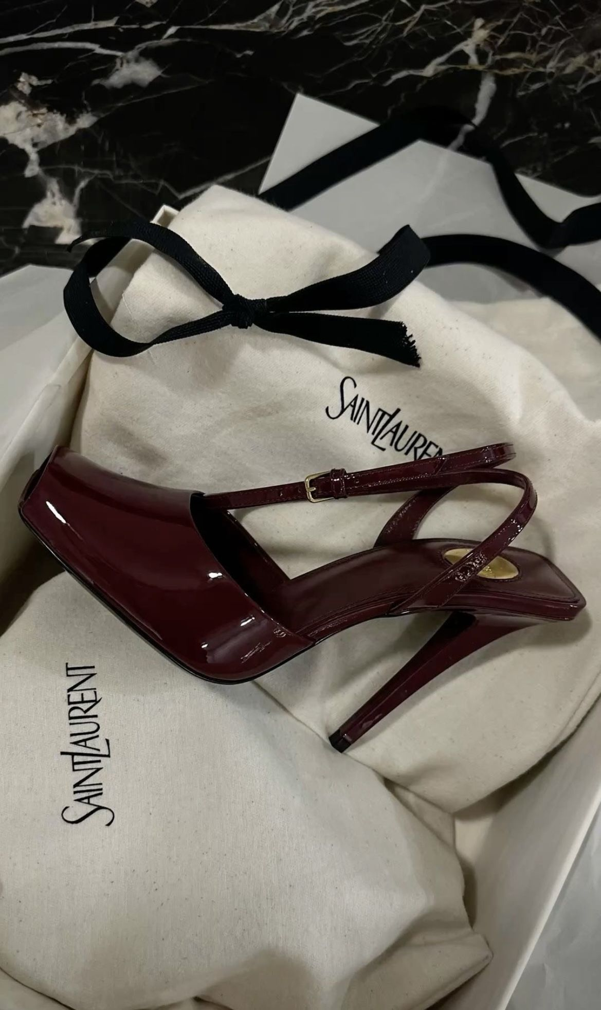 A pair of dark red high-heeled shoes with a cross strap, placed on a white Saint Laurent shoe box and dust bag with black branding, with a black fabric ribbon tied into a bow.