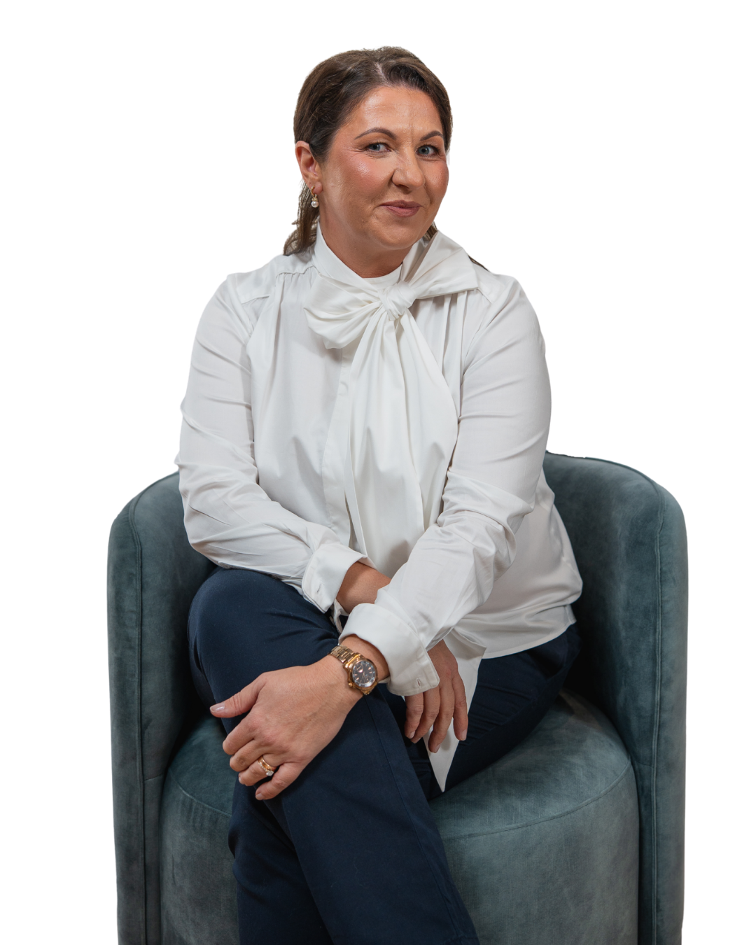 Magda Campbell with brown hair styled to the side, wearing a white blouse with a bow tie at the neck, dark blue pants, a watch, and earrings, sitting on a gray velvet armchair with her arms crossed and smiling at the camera.