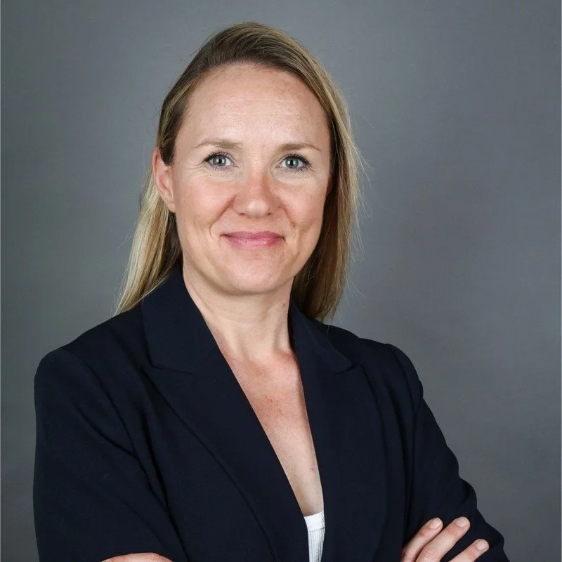 Professional woman with blonde hair wearing a dark blazer, standing against a gray background.