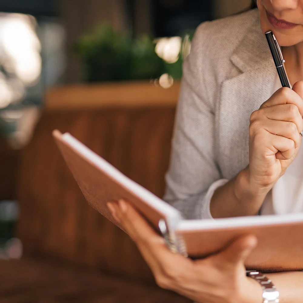 A woman holding a pen and a notebook while sitting at a table in a cozy indoor space.