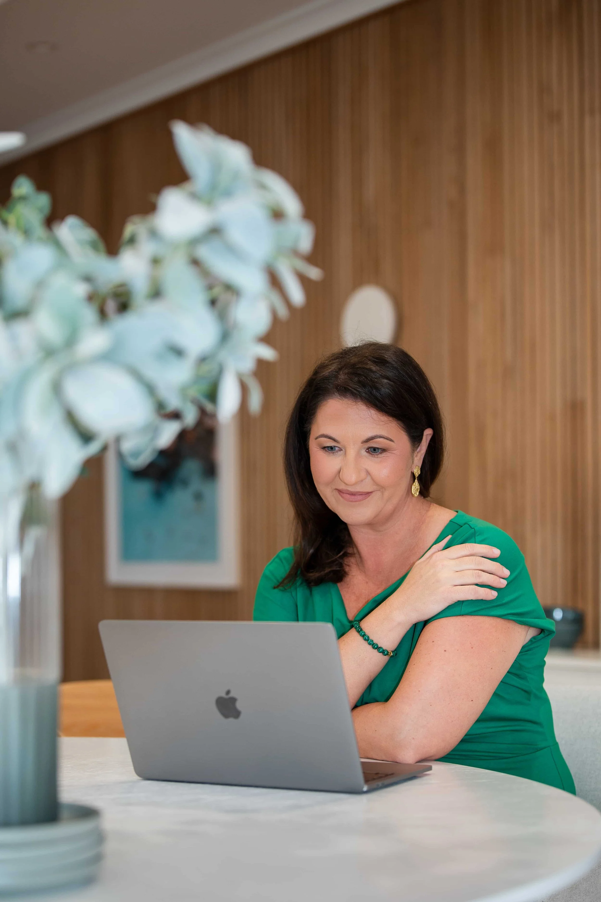 A woman in a green dress sitting at a desk with a silver MacBook laptop, touching her shoulder and looking at the screen. A vase with white flowers is in the foreground, slightly out of focus, and wooden paneling is on the wall behind her.