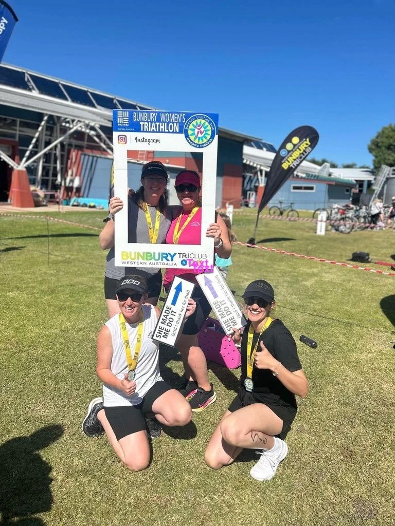 Magda Campbell and friends celebrating after a triathlon, holding a large frame with event branding, medals around their necks, and signs with motivational messages. They are outdoors on grass with event buildings, and flags in the background.