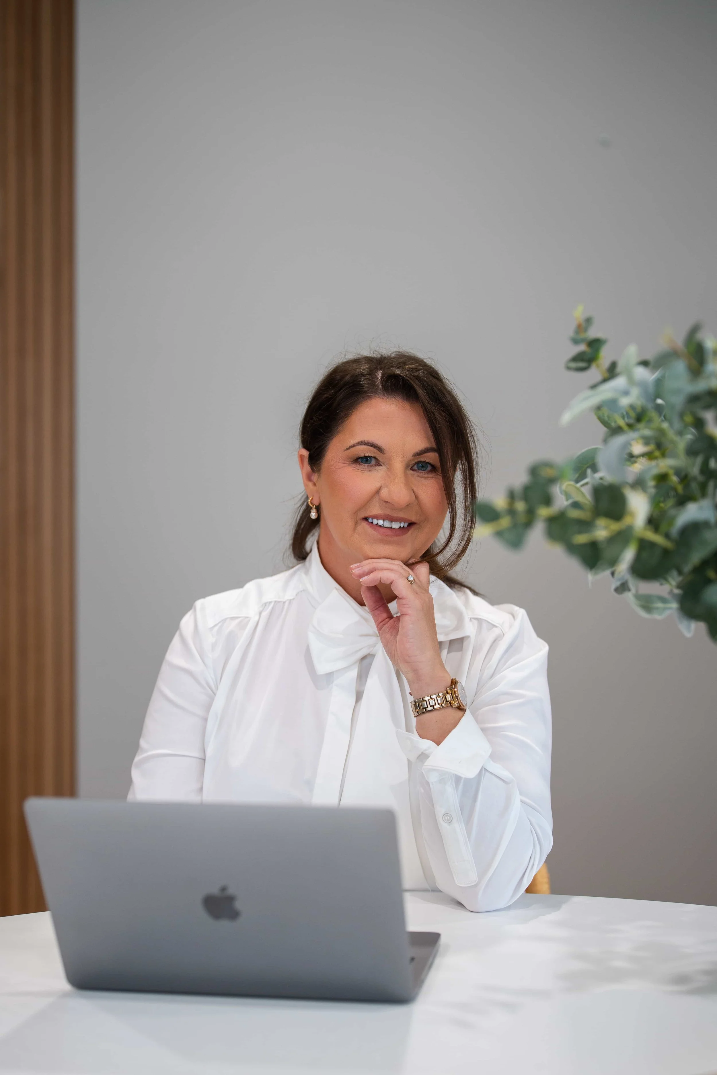 Magda Campbell sitting at a white desk, smiling, with a silver MacBook in front of her. She is wearing a white shirt, gold earrings, and a gold watch, and is looking into a mirror, with a large green plant visible to her right.