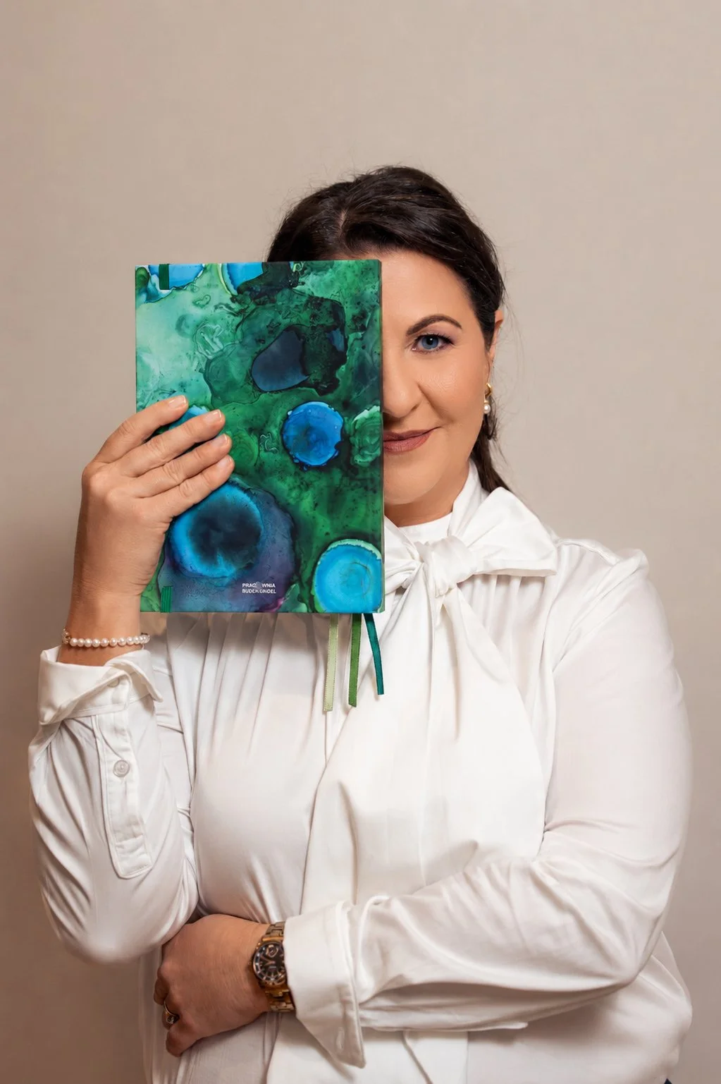 Magda Campbell wearing a white blouse with a bow, holds a colorful abstract art book in front of her face, covering half of her face, standing against a plain beige wall.