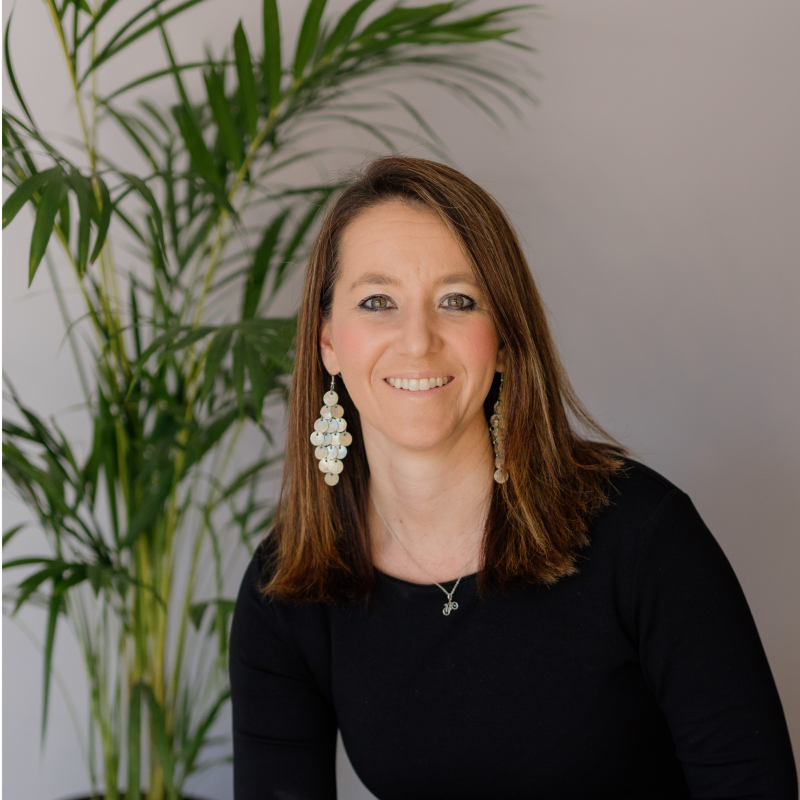 A smiling woman with shoulder-length brown hair, wearing a black top and large dangling earrings, sitting in front of a green potted plant on a white wall background.