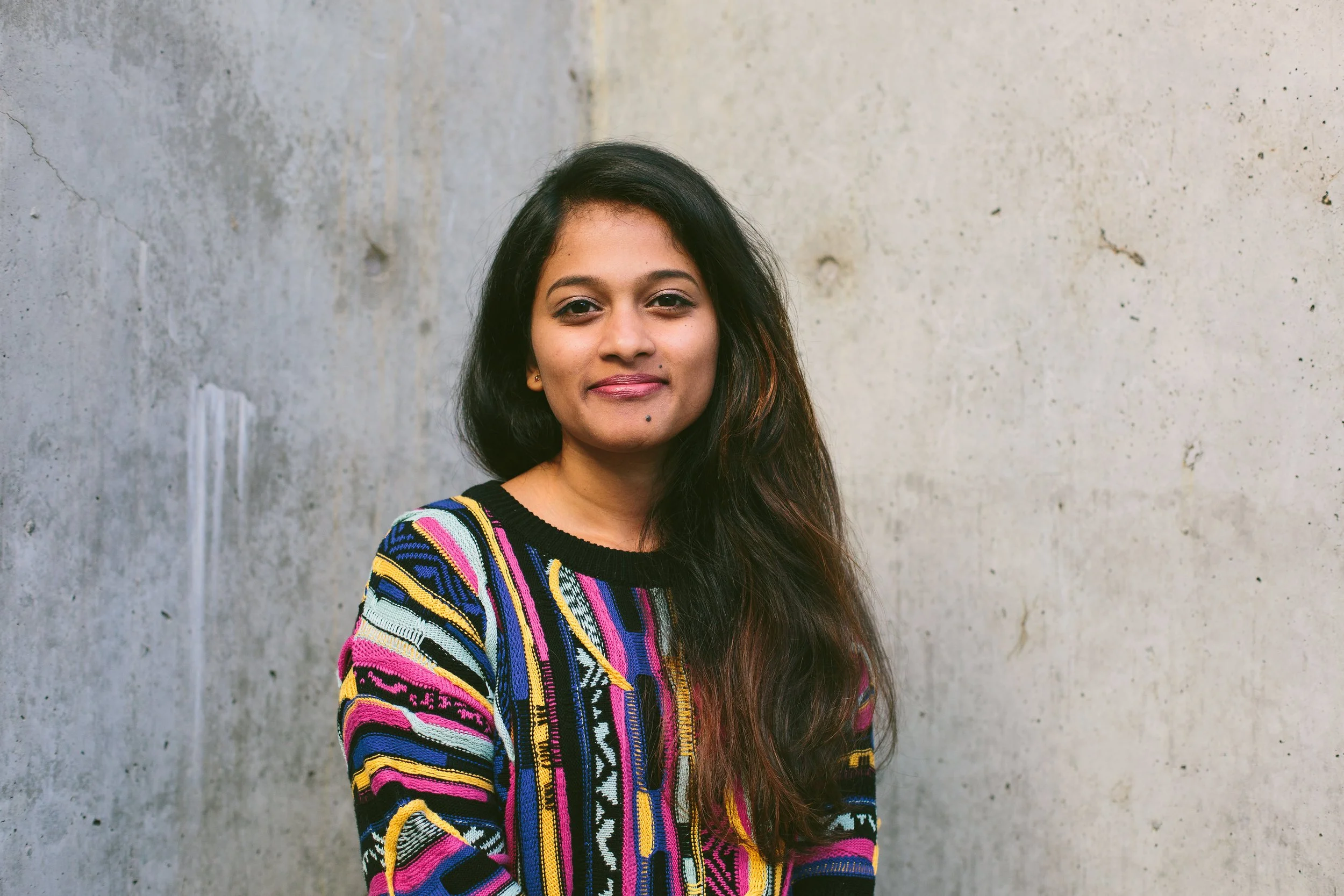 A young woman with long dark hair standing against a plain concrete wall, wearing a colorful striped sweater and smiling subtly.