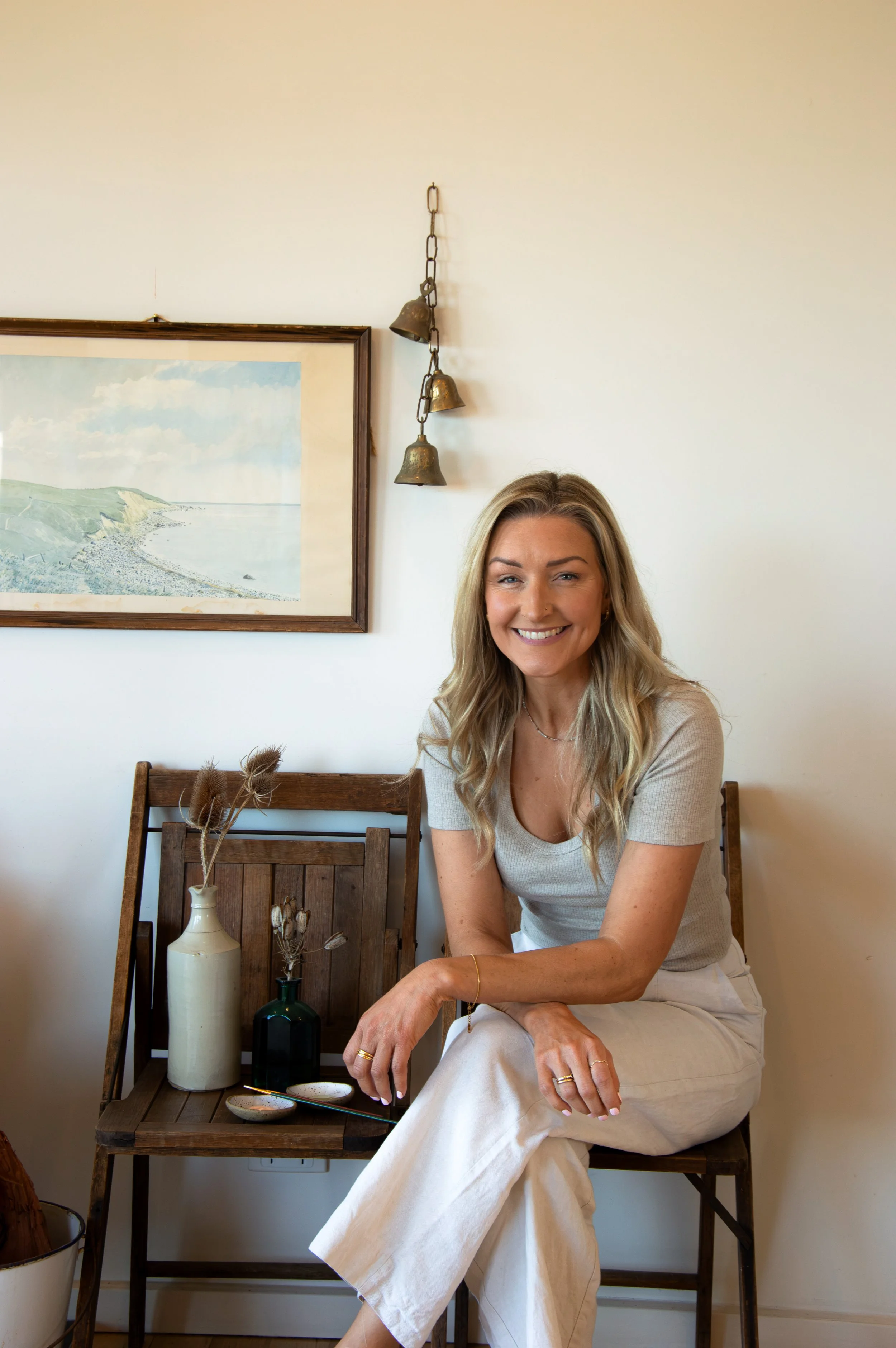 A woman with long blonde hair smiling and sitting on a wooden chair in a room with beige walls. Behind her is a framed landscape painting and a wall decoration with three small metal bells hanging from a chain. She is wearing a light gray short-sleeve top and beige pants, with jewelry on her fingers and wrist. There are decorative vases with dried plants on a wooden shelf in front of her.