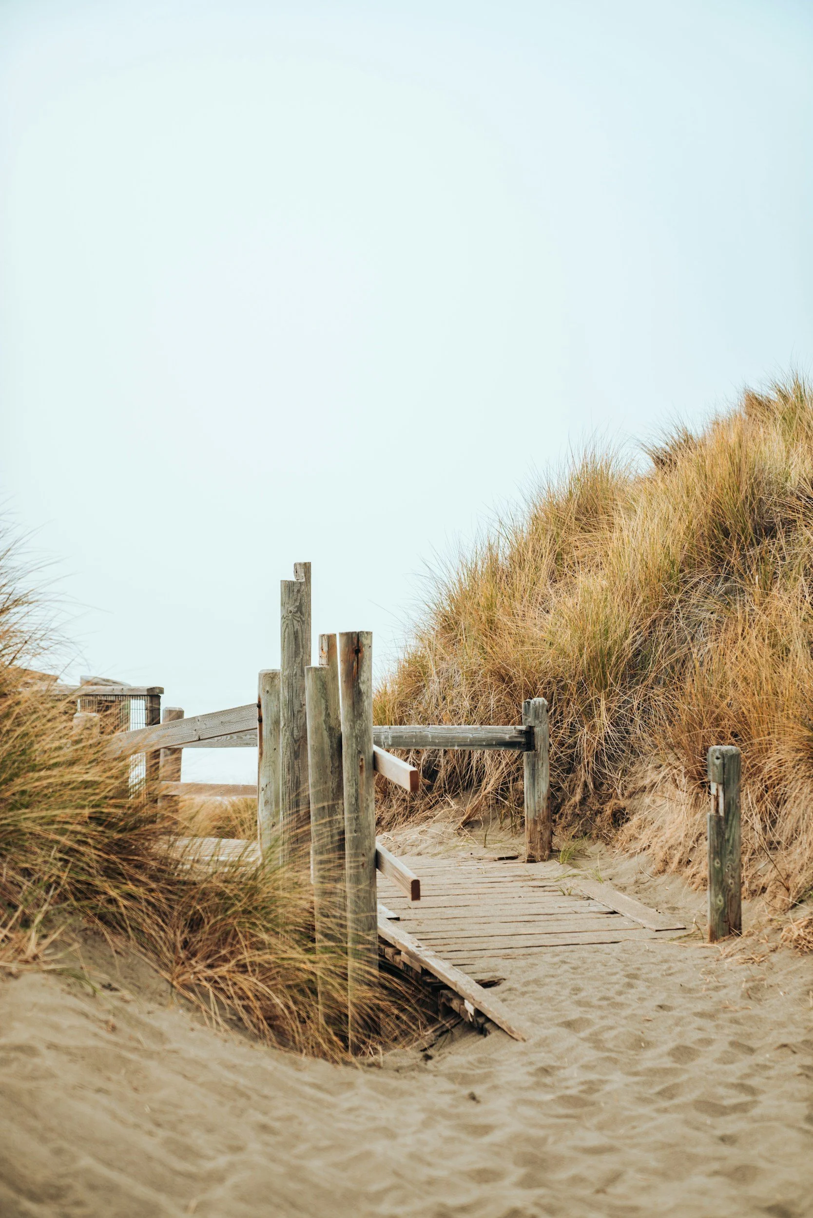A sandy beach with a wooden boardwalk leading through tall grass dunes, with a partly cloudy sky.