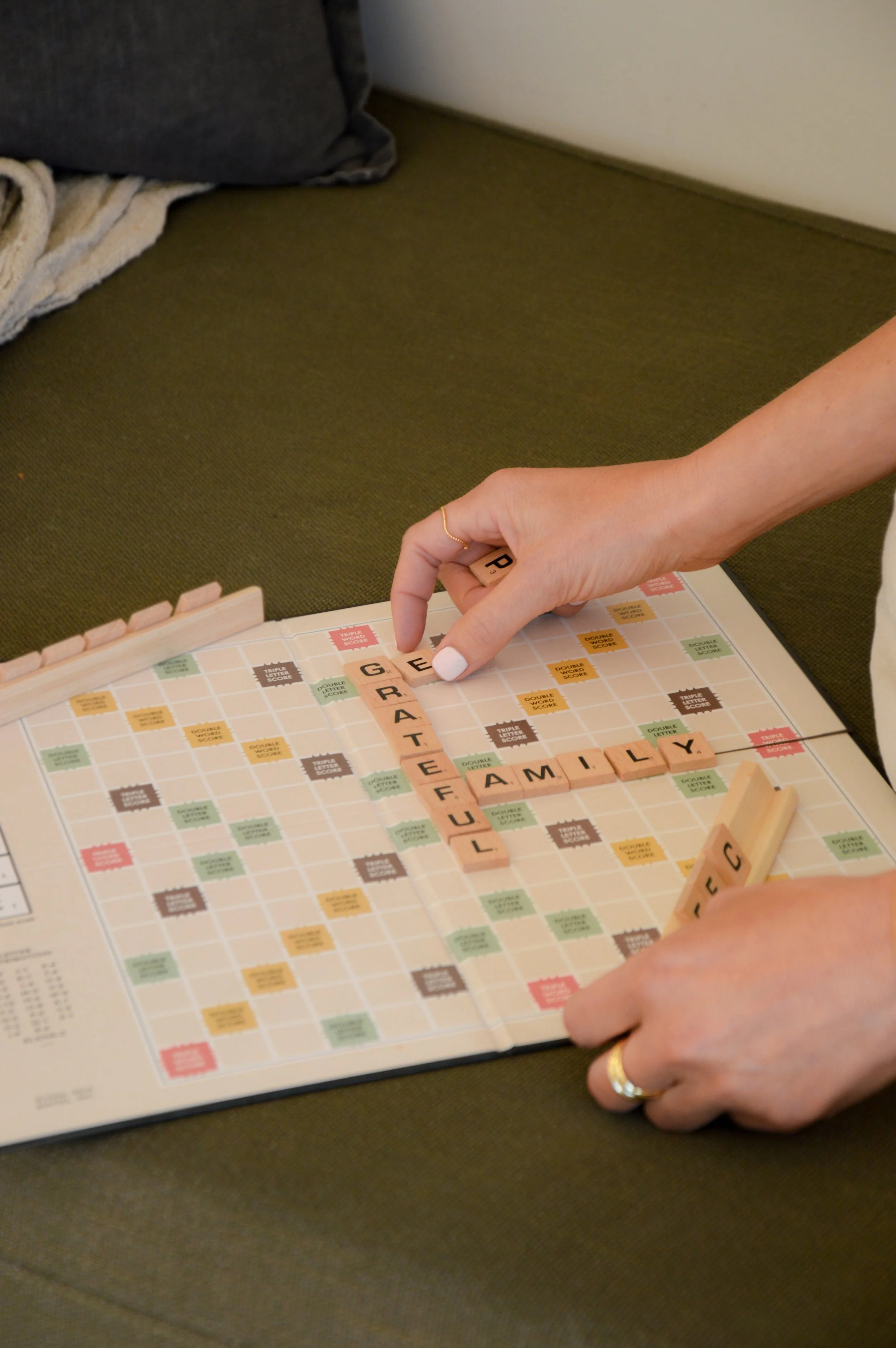 Close-up of a person's hand with a gold ring, playing Scrabble on a board with tiles spelling 'GREAT FAMILY,' on a table with a dark surface, with a couch and blanket in the background.
