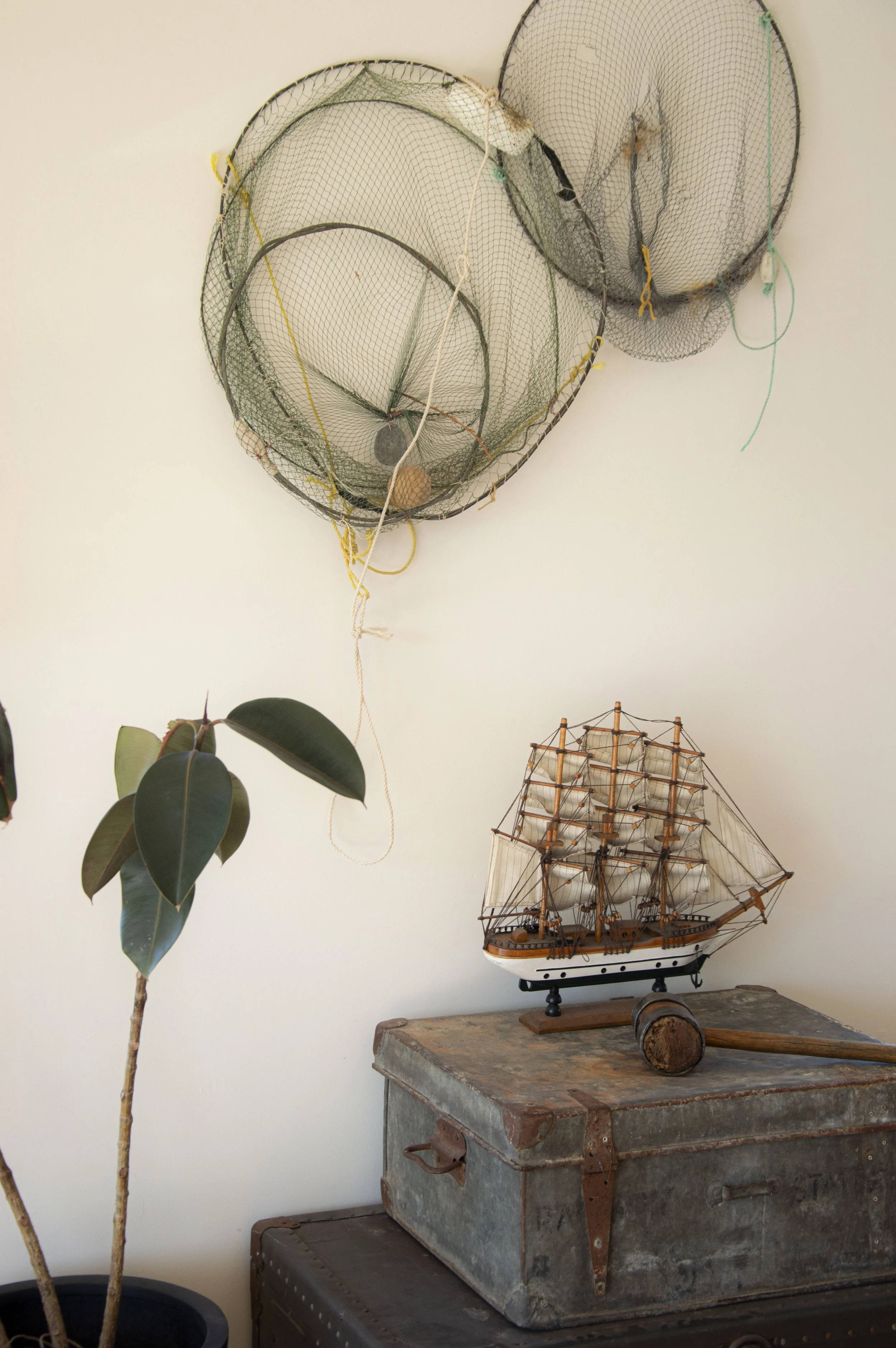 A wall with hanging fishing nets, a potted plant with dark green leaves, and a model ship on a wooden table with a vintage metal trunk underneath.
