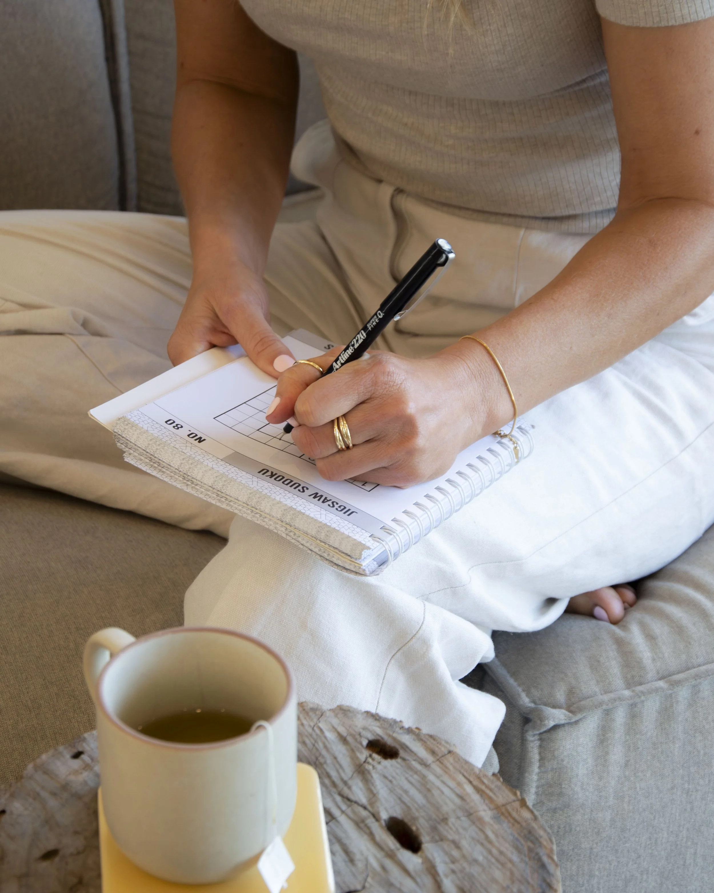 Person sitting on a cushioned seat, writing on a notepad with a black pen, with a cup of coffee or tea on a wooden table in front.