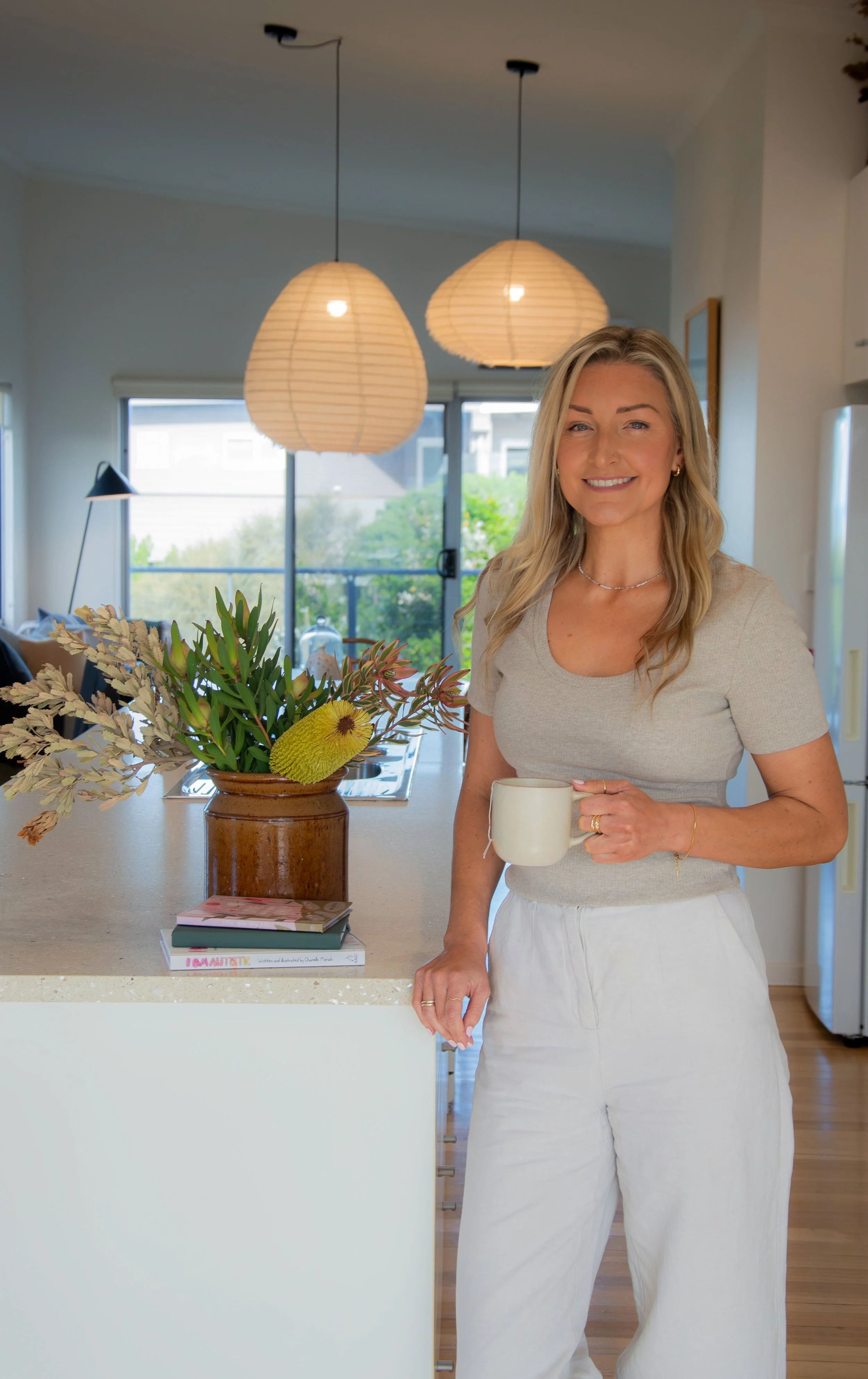 A woman standing in a kitchen, holding a white mug, smiling, with a bouquet of flowers on the counter in front of her, and pendant lights hanging from the ceiling in the background.