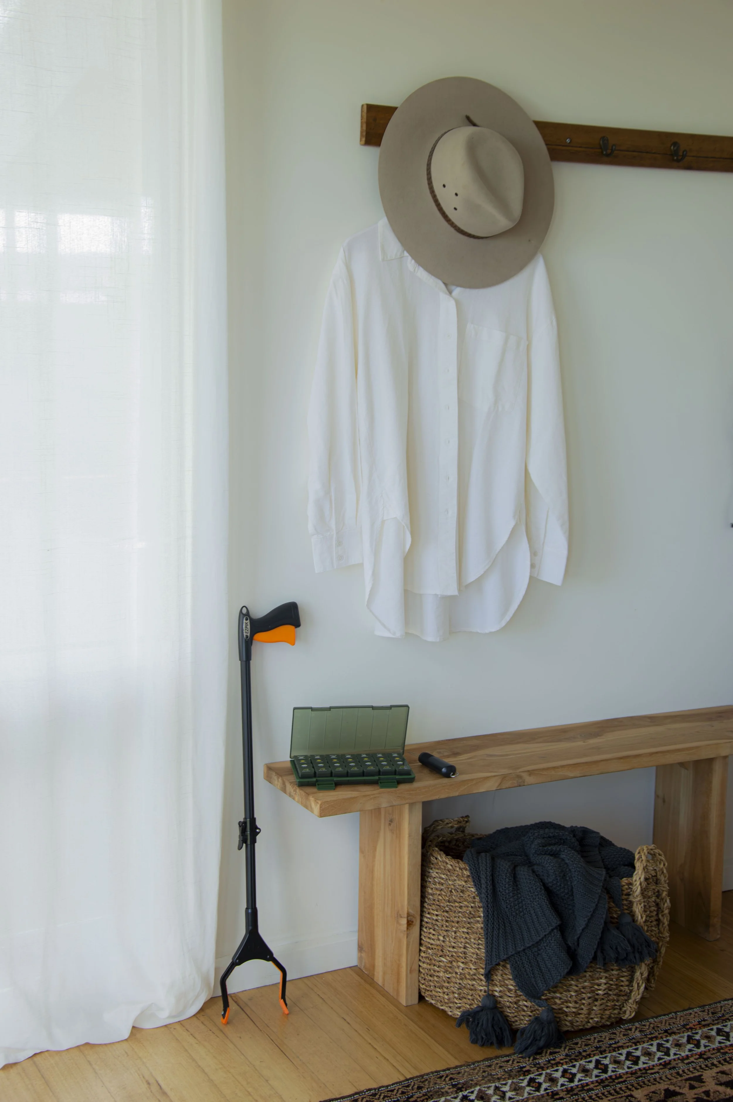 A wooden table with a woven basket filled with black clothing, a black flashlight, and a green plastic container with a latch. A white shirt hangs from a wall hook topped by a wide-brimmed beige hat. A black rifle-like object leans against the wall, 