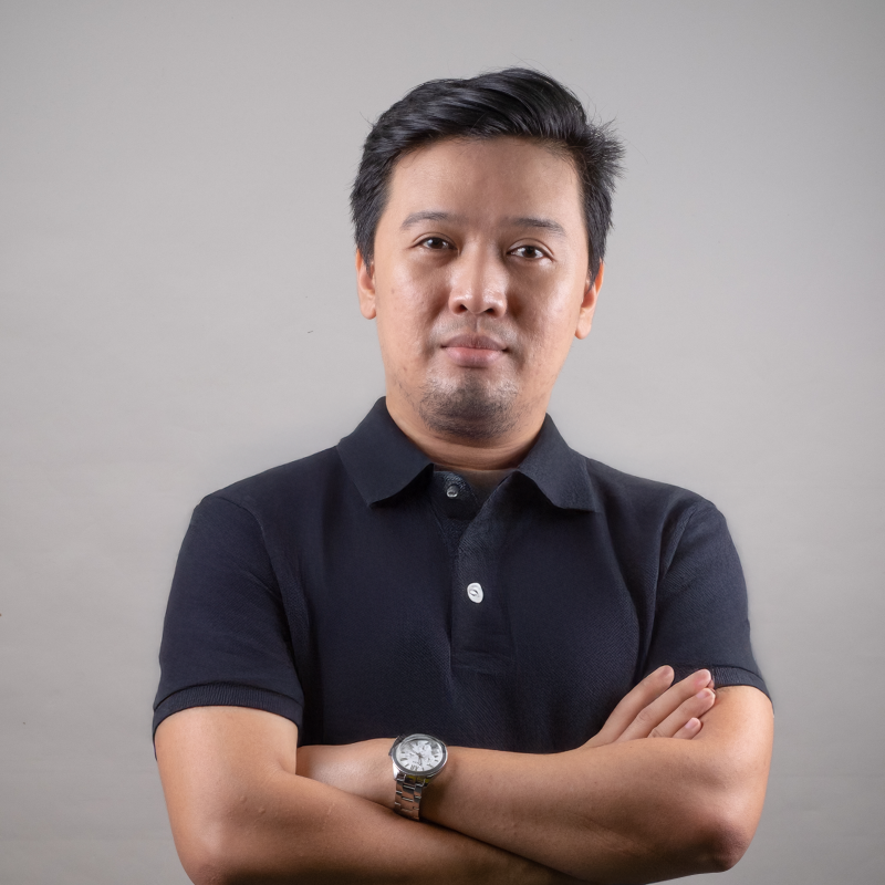 Portrait of a man with crossed arms wearing a black polo shirt and watch, standing against a plain gray background.