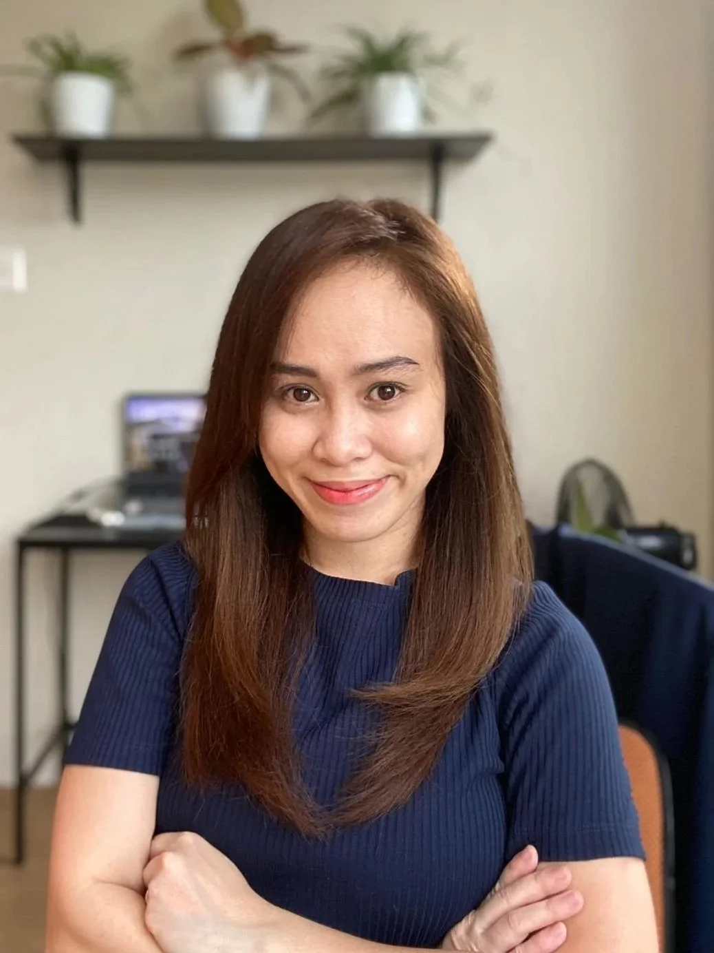 A woman with shoulder-length brown hair and a blue top smiling with arms crossed in an office setting with a shelf holding potted plants in the background.